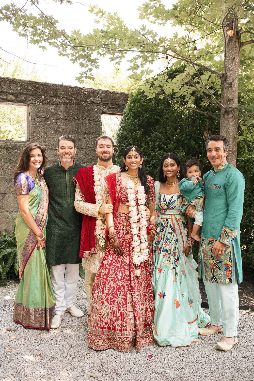 Group of seven people, including a bride and groom in traditional Indian attire with flower garlands, posing outdoors among trees and greenery.