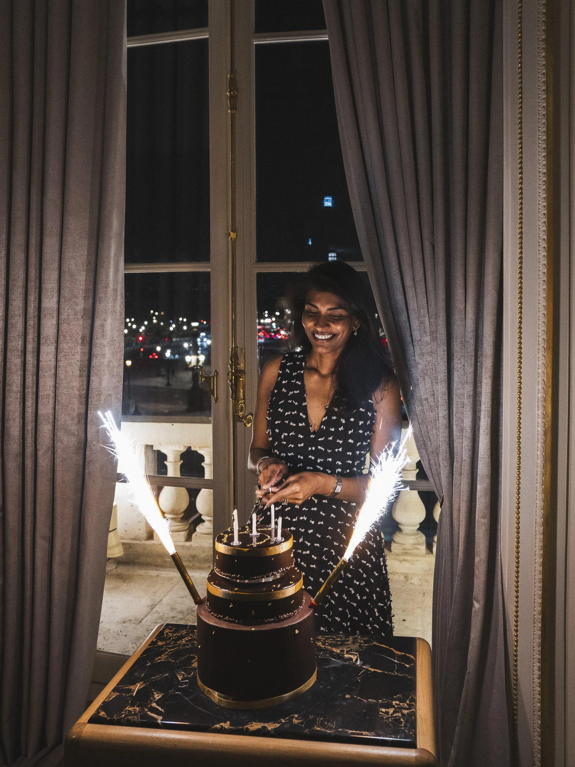 A woman in a black and white dress celebrating with a birthday cake and sparklers by a window at night.