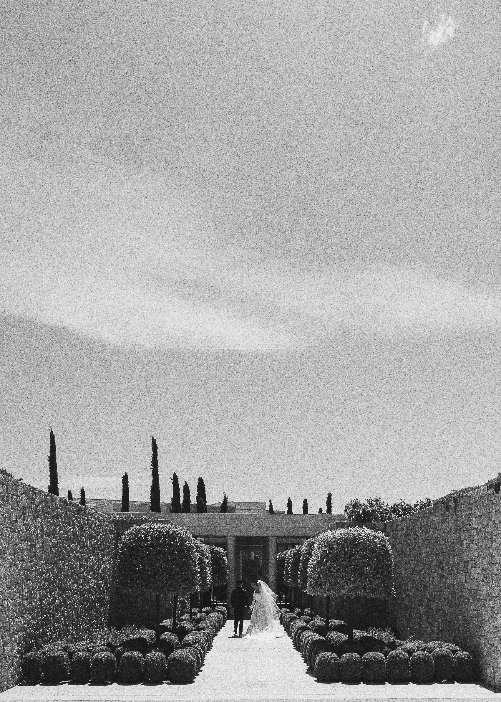 A bride and groom walking hand in hand down a garden aisle lined with rounded bushes, with stone walls and tall cypress trees on either side, beneath a partly cloudy sky.