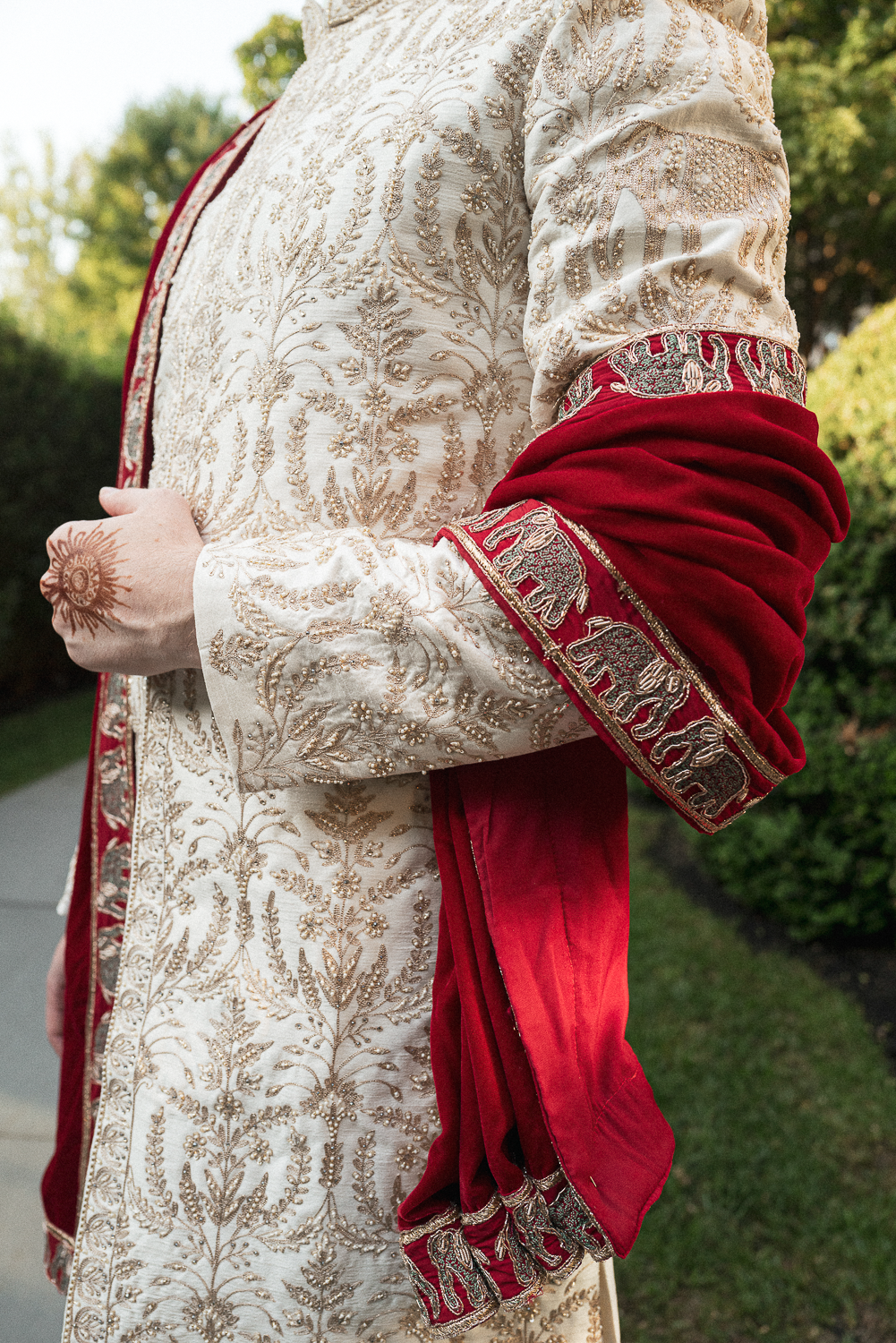 Close-up of a person wearing a cream-colored traditional outfit with gold embroidery and a red velvet shawl with intricate embroidery, standing outdoors.