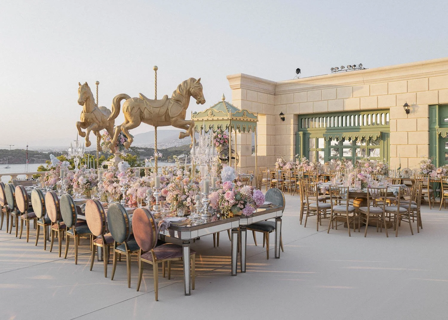 Elegant outdoor event space with pink and white floral centerpieces, vintage chairs, and carousel horse sculptures on a terrace with city and water views at sunset.