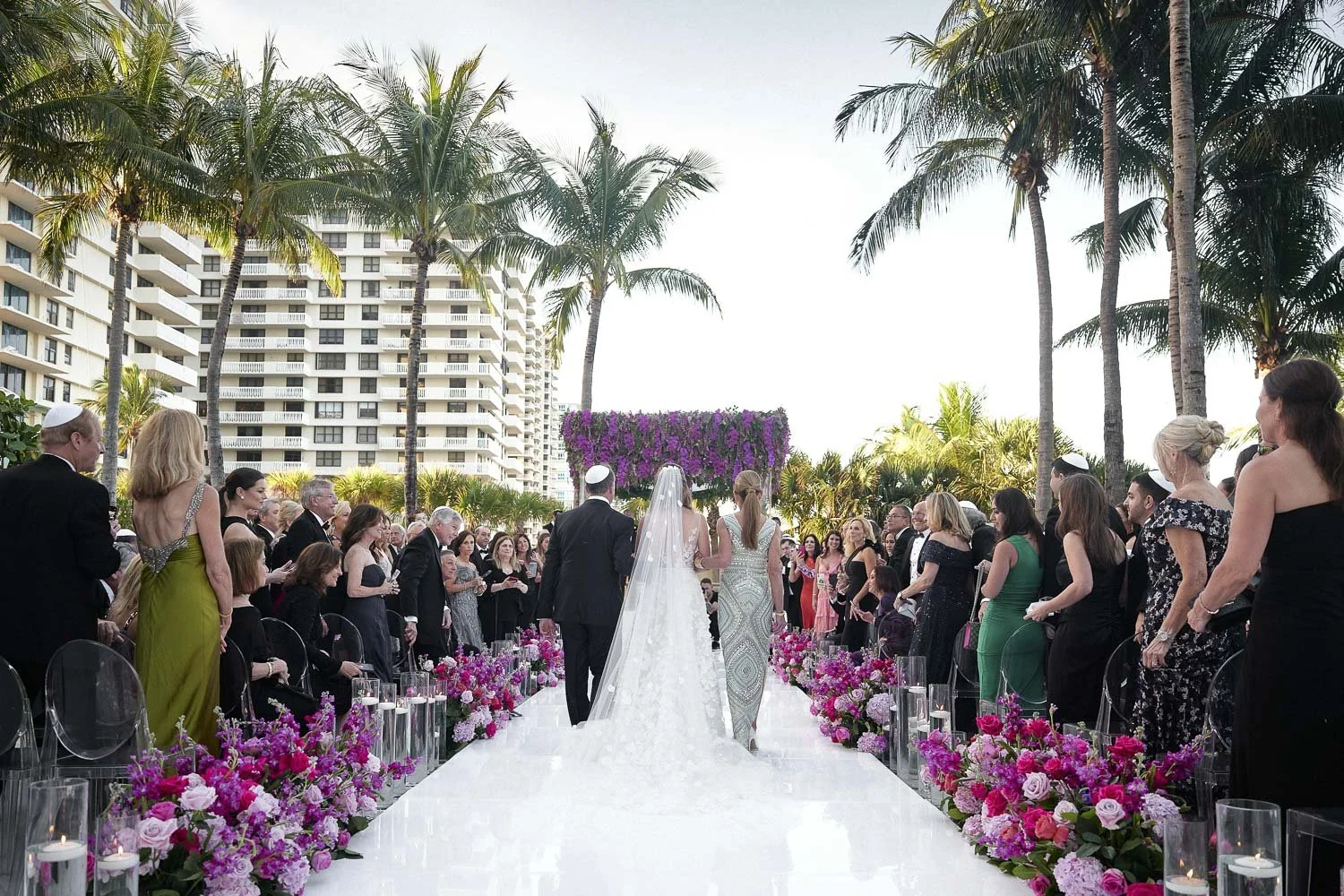 Wedding ceremony outdoors on a white aisle decorated with pink and purple flowers, surrounded by guests dressed in formal attire, with tall palm trees and high-rise buildings in the background.