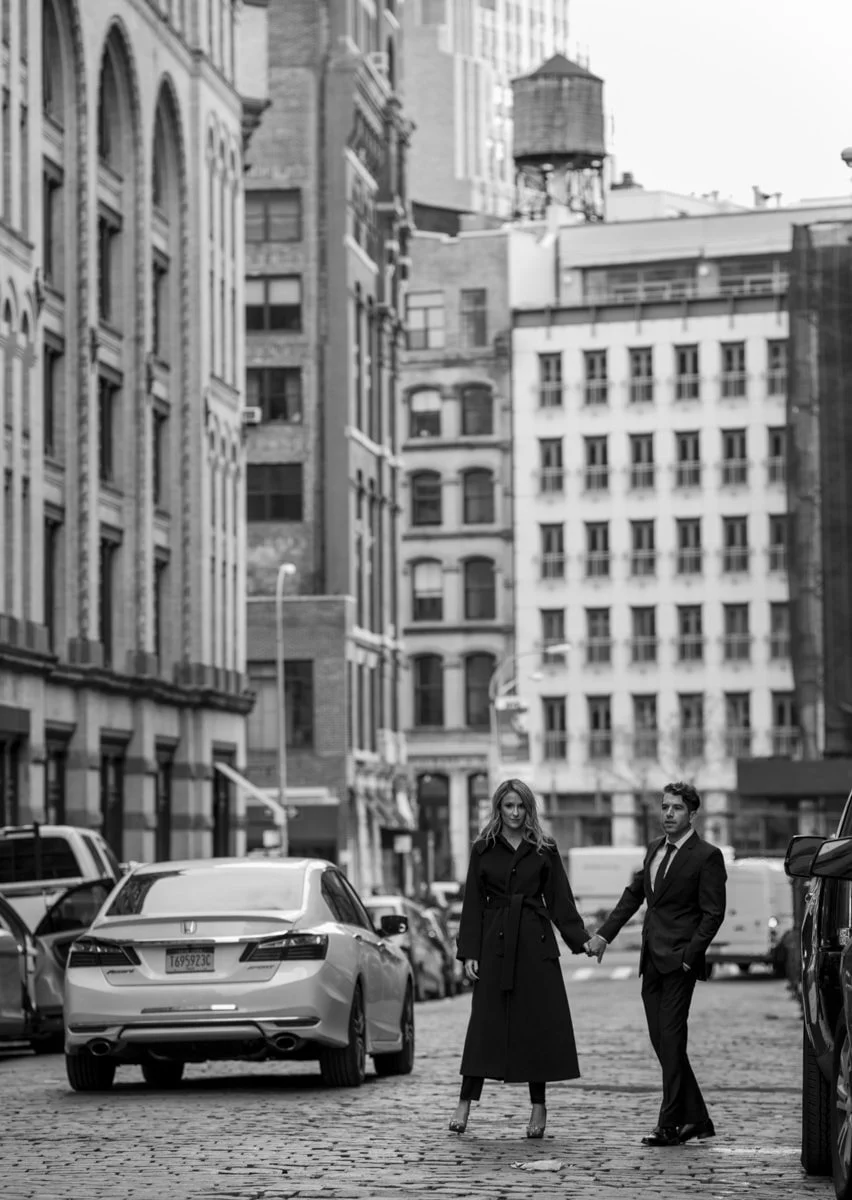 A black and white photo of a woman and a man holding hands, walking on a cobblestone street in an urban city with tall buildings around.