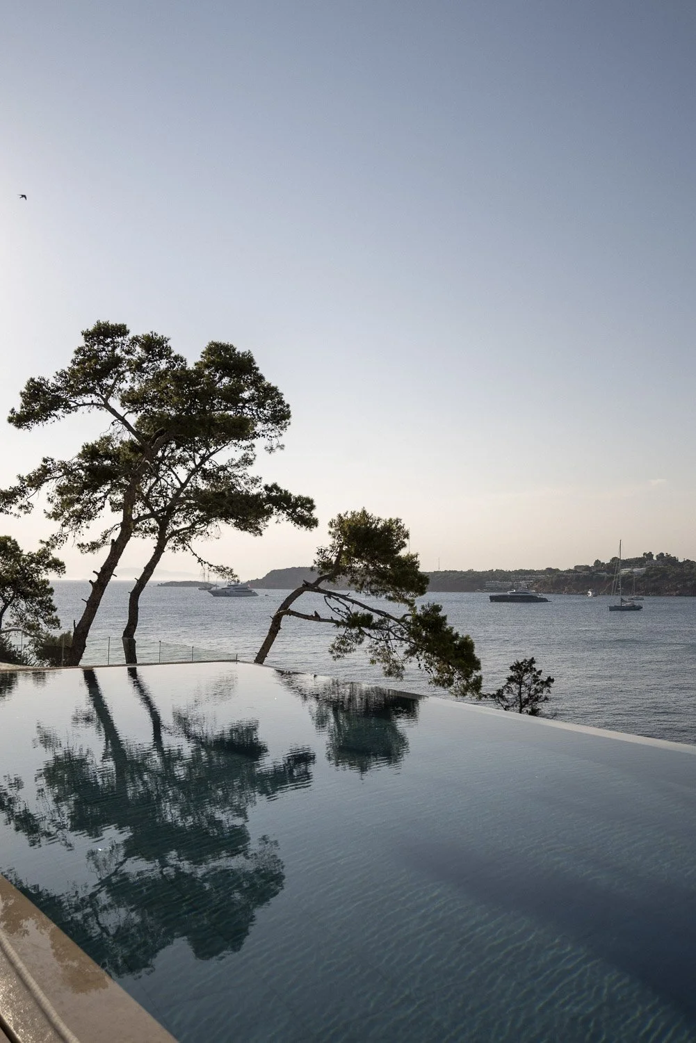 A view of a swimming pool with reflections of trees, overlooking the ocean with sailboats and yachts, under a clear sky.
