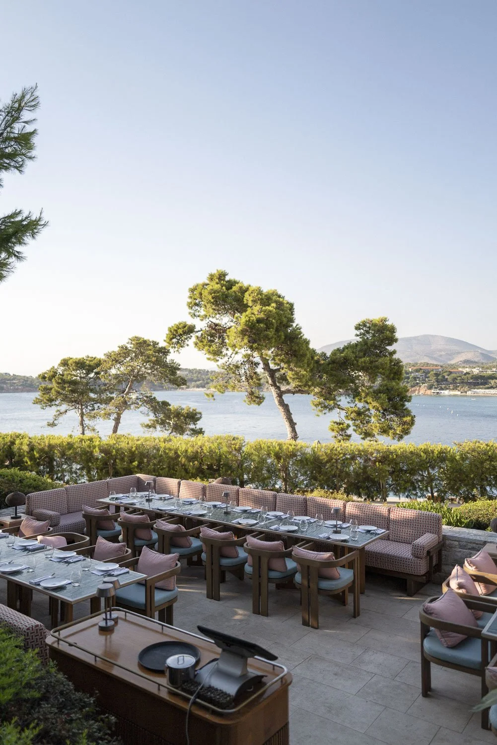 Outdoor dining area with tables set for a meal, overlooking a body of water with trees and hills in the background.