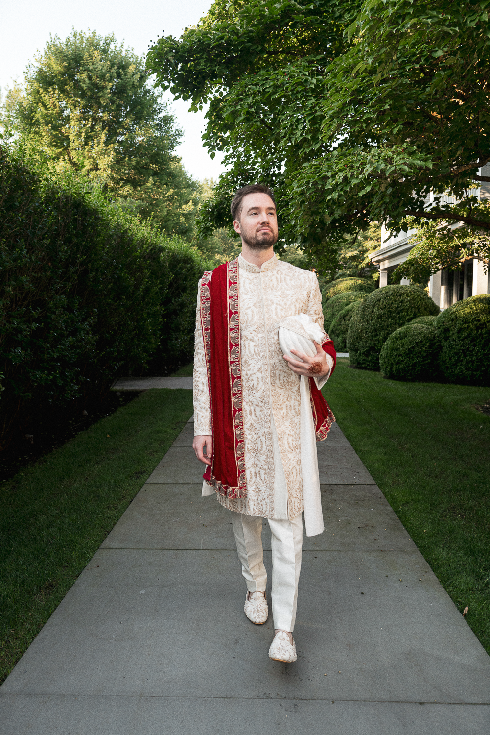 A man dressed in traditional Indian wedding attire walking on a sidewalk in a garden.