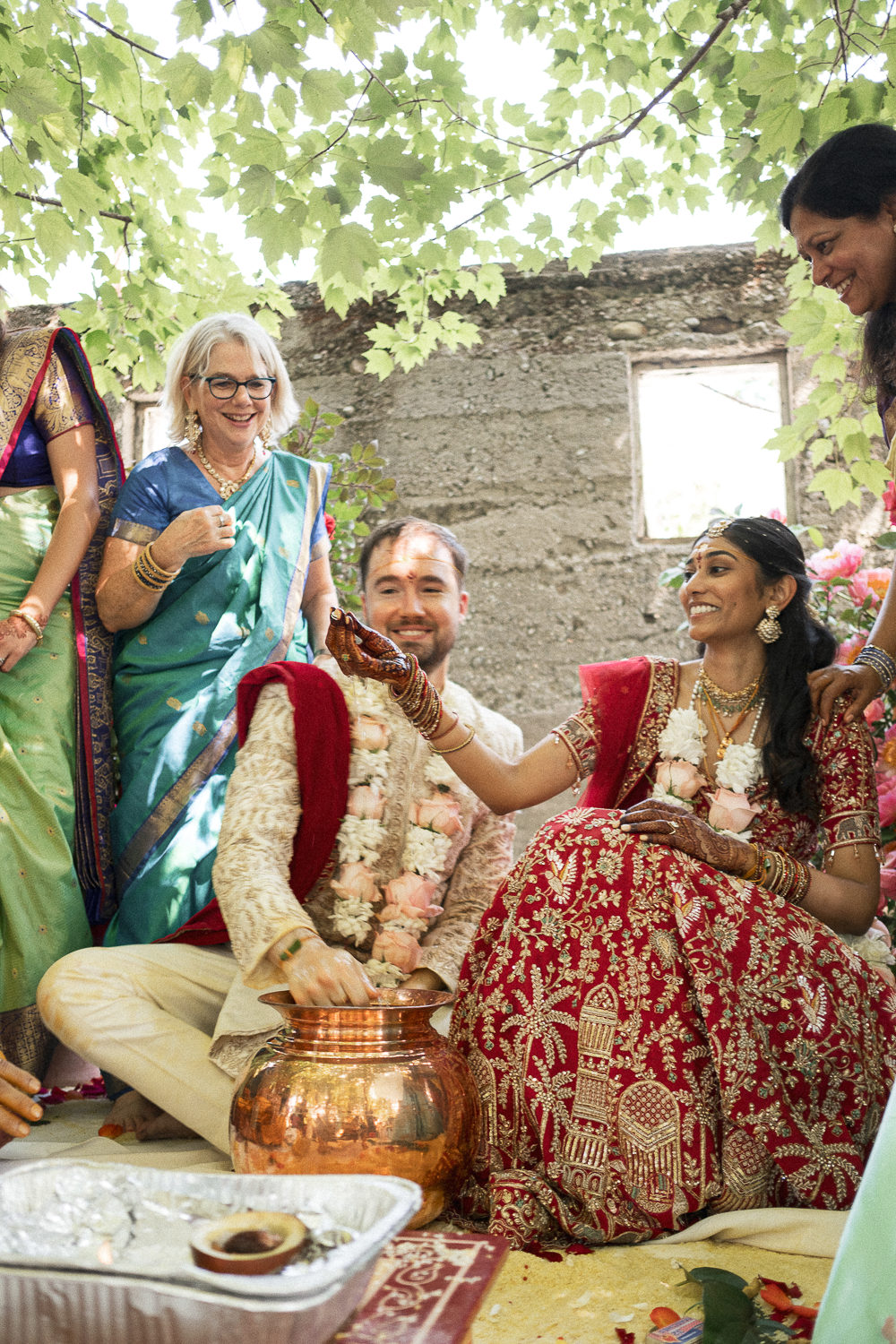 Indian wedding ceremony with a bride and groom in traditional attire, surrounded by women in colorful sarees, with leaves overhead and a rustic wall in the background.