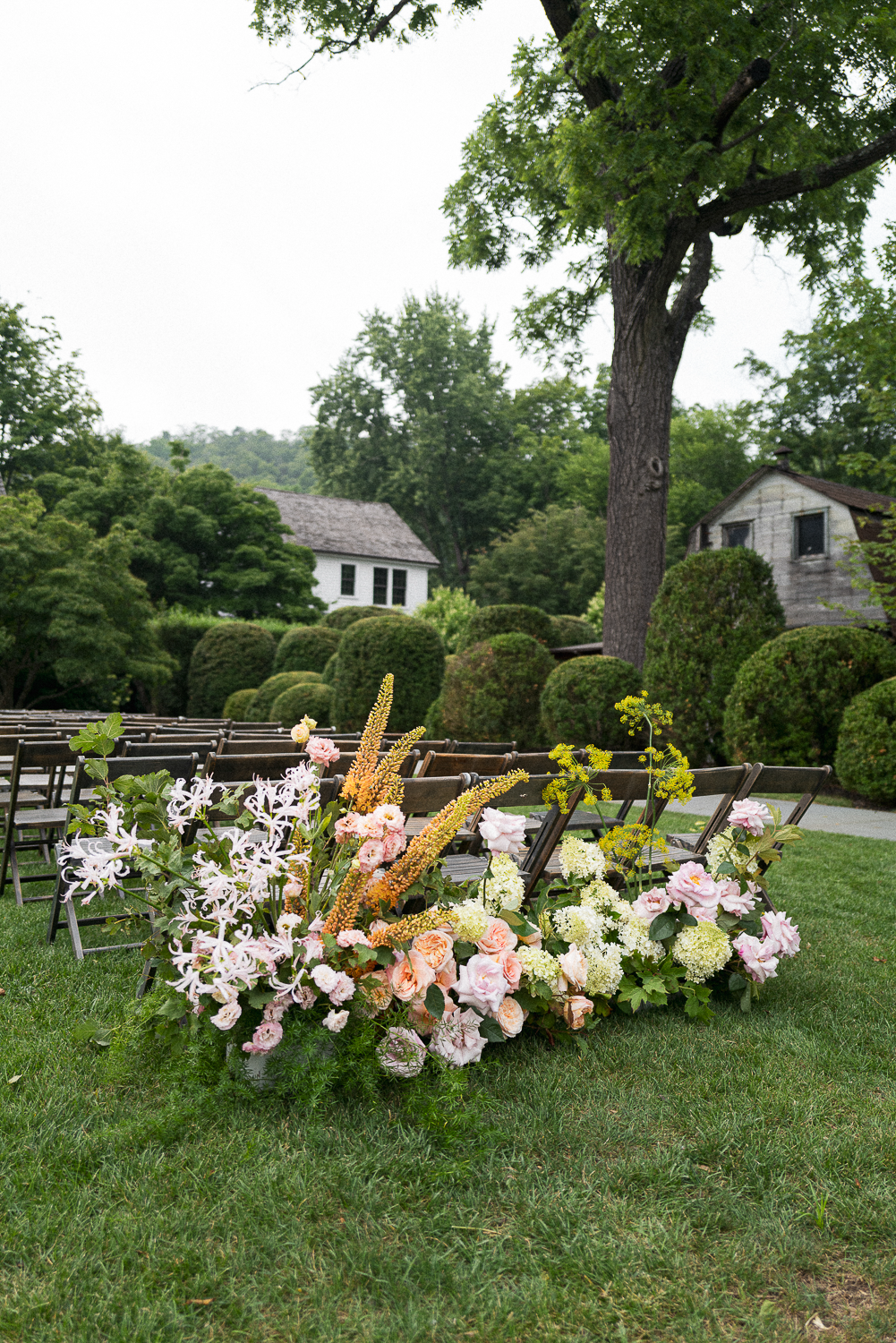 Flower arrangements with pink and white flowers, set up outdoors for a wedding or event, with green grass, trees, and houses in the background.