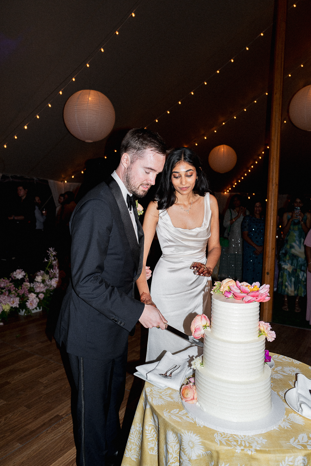 A bride and groom cutting a wedding cake at their reception under hanging lanterns and string lights.