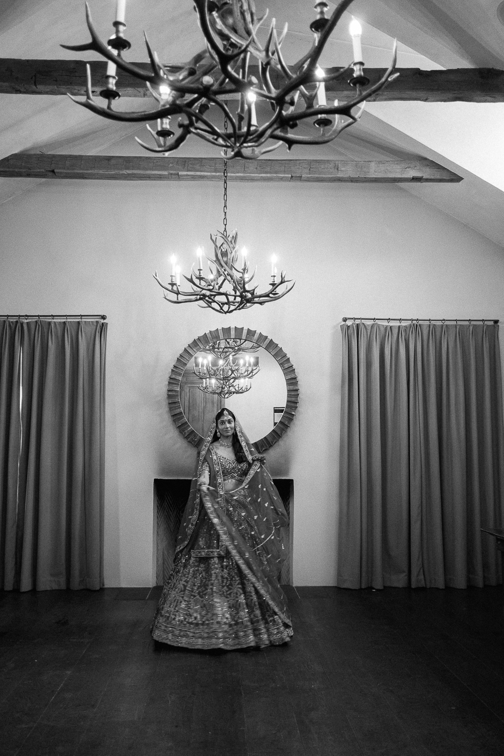 A woman dressed in traditional Indian bridal attire standing in a room with curtains, a large circular mirror on the wall, and two chandeliers hanging from the ceiling.