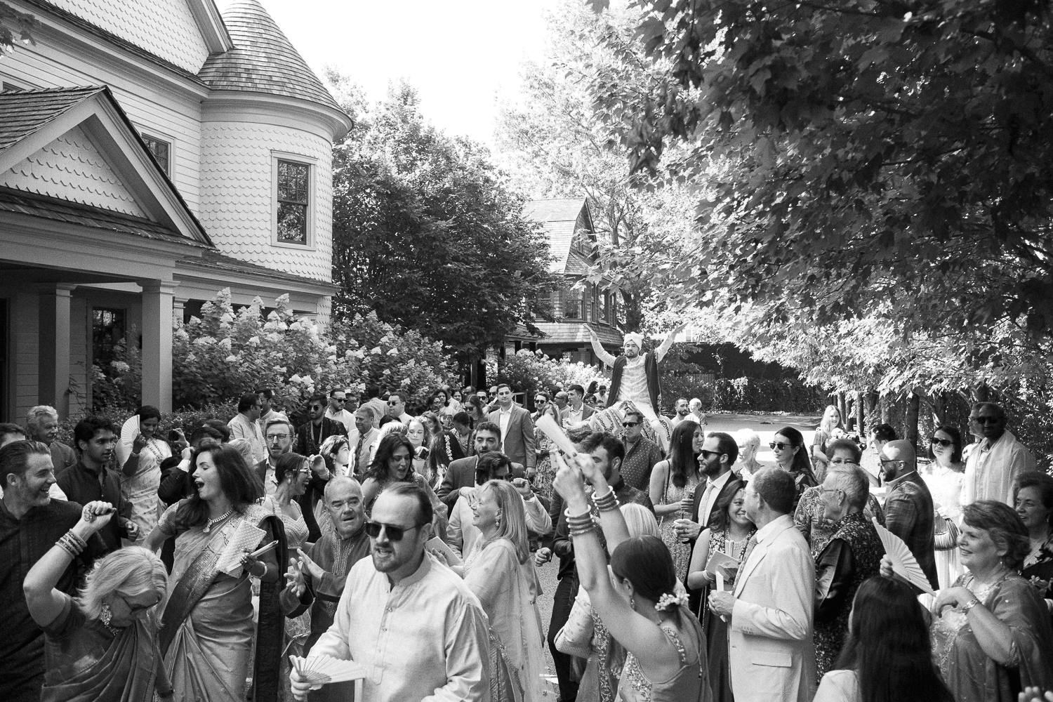 A black and white photo of a diverse crowd of people gathered outside a large girl in traditional clothing and others in formal attire. The group appears to be celebrating, with some people smiling, dancing, and holding fans. The setting is a residen