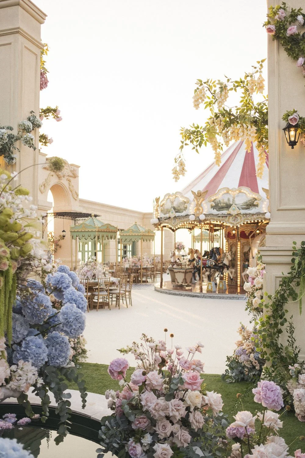 Decorative scene at a vintage-style fairground with a carousel, flower arrangements, outdoor seating, and pastel-colored structures, bathed in soft daylight.