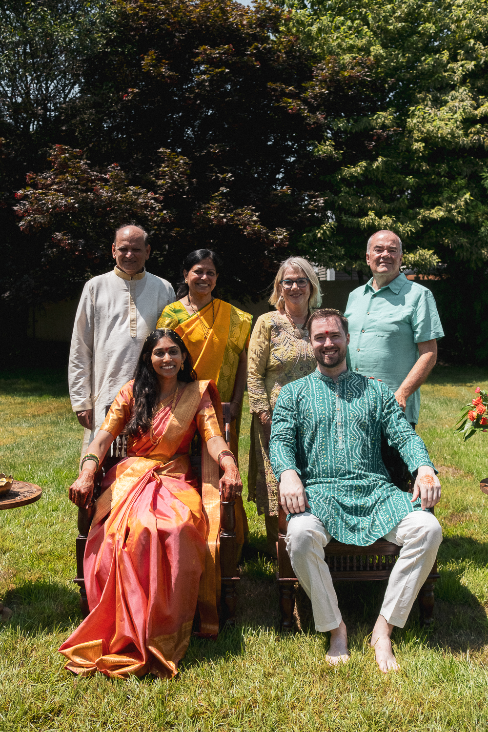 Group of six people of diverse backgrounds, dressed in traditional Indian attire, sitting and standing outdoors on a sunny day with lush green trees in the background.