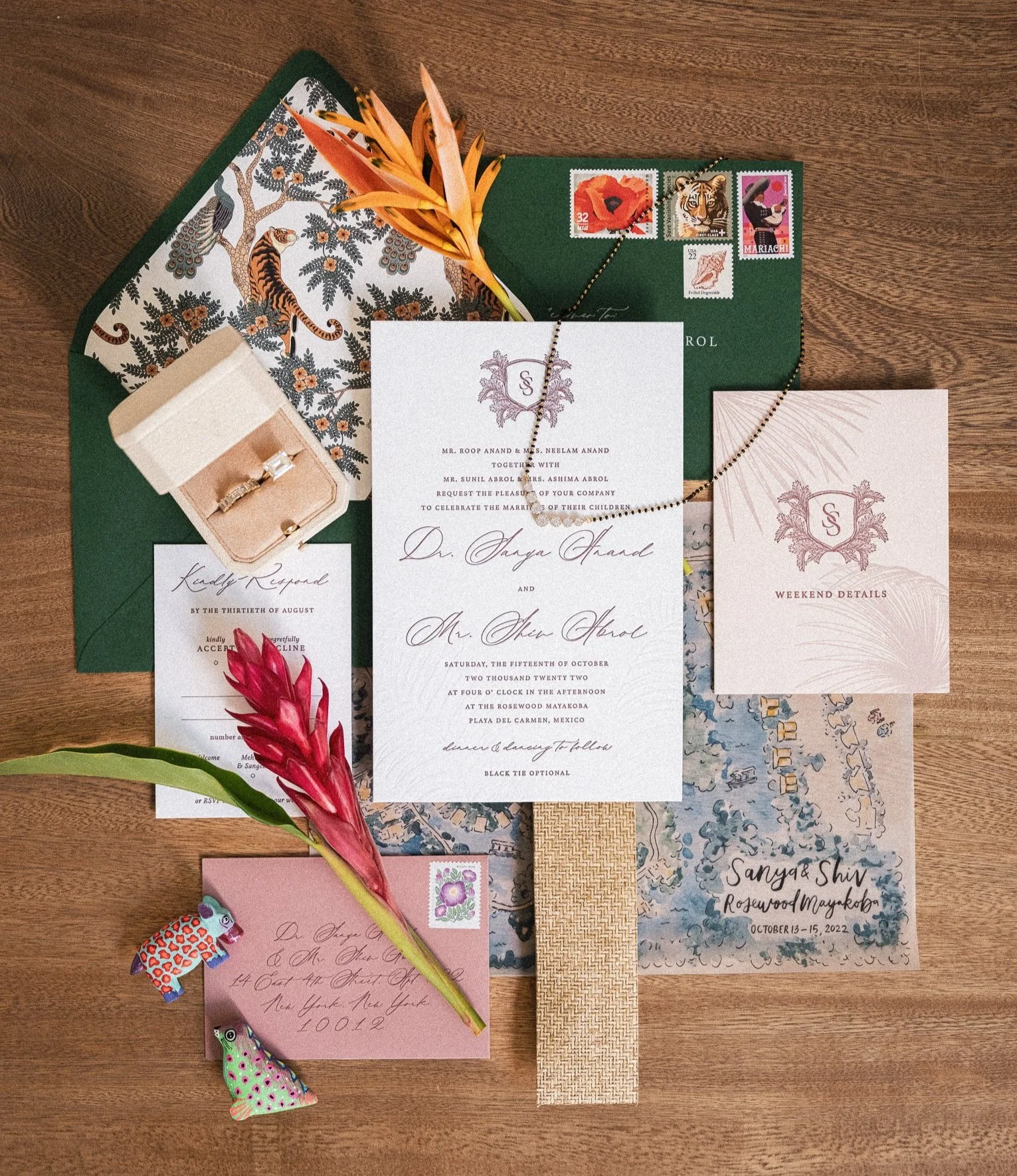 A collection of wedding invitations, stamps, a small ring box with a ring, a beaded necklace, a red flower, and a small colorful stuffed animal on a wooden surface.