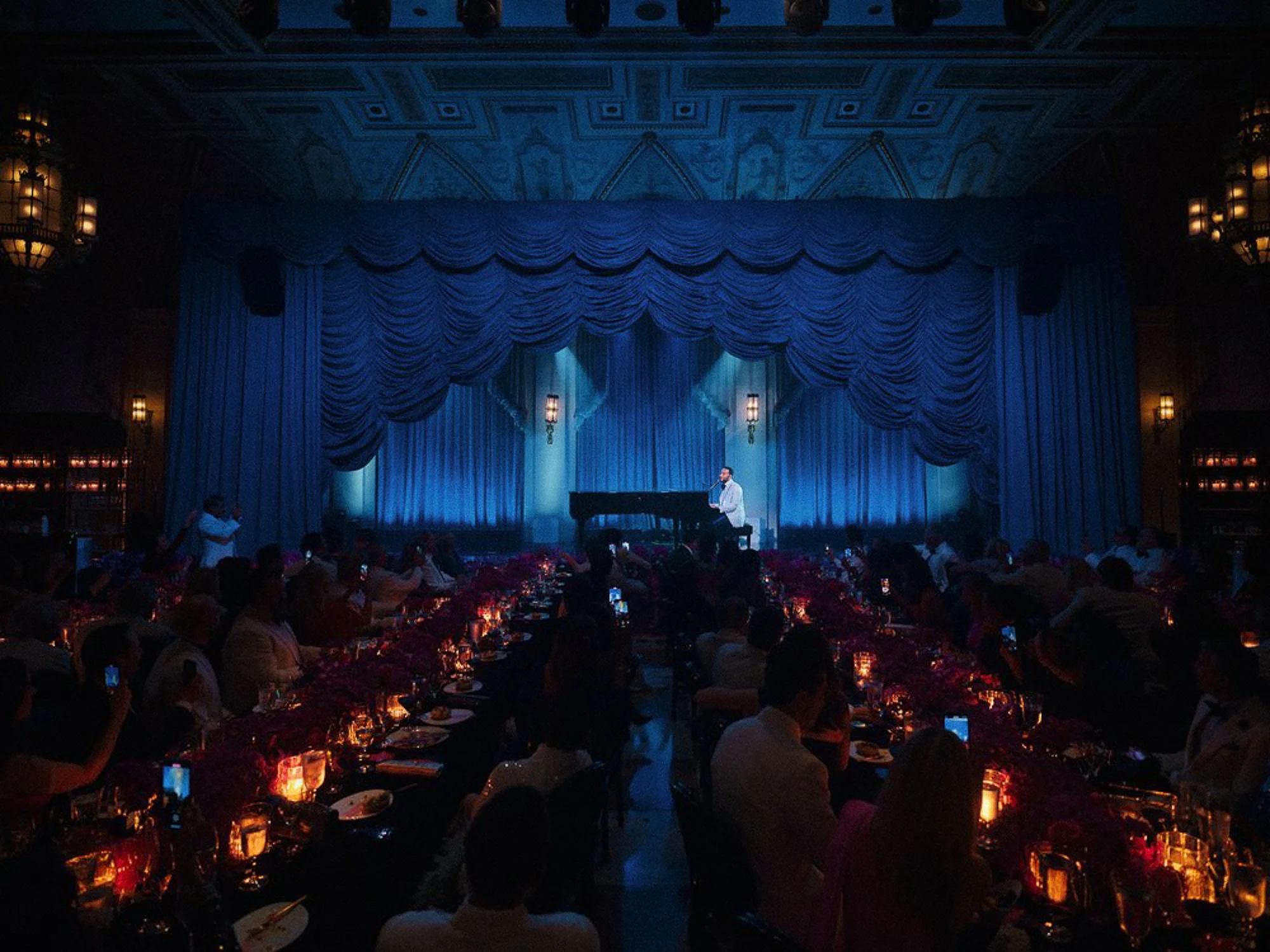 A performer in a white shirt and dark pants standing at a microphone on a stage with blue curtains and ornate blue ceiling. Audience members are seated at tables with candles and floral centerpieces, watching the performance in a dimly-lit theater or ballroom.