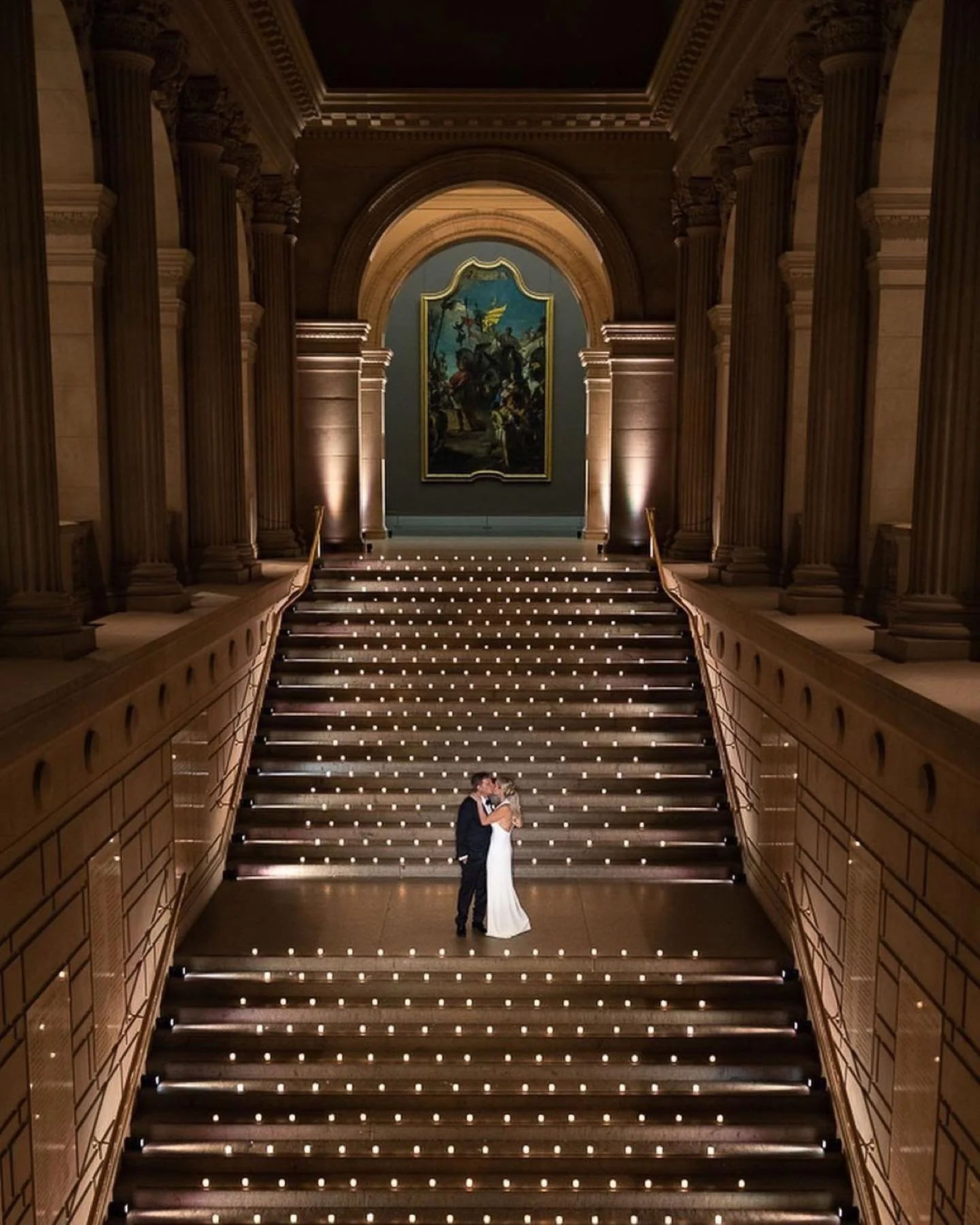 A couple dressed in wedding attire dancing on a grand staircase inside a luxurious building with ornate columns and a large portrait hanging at the top.
