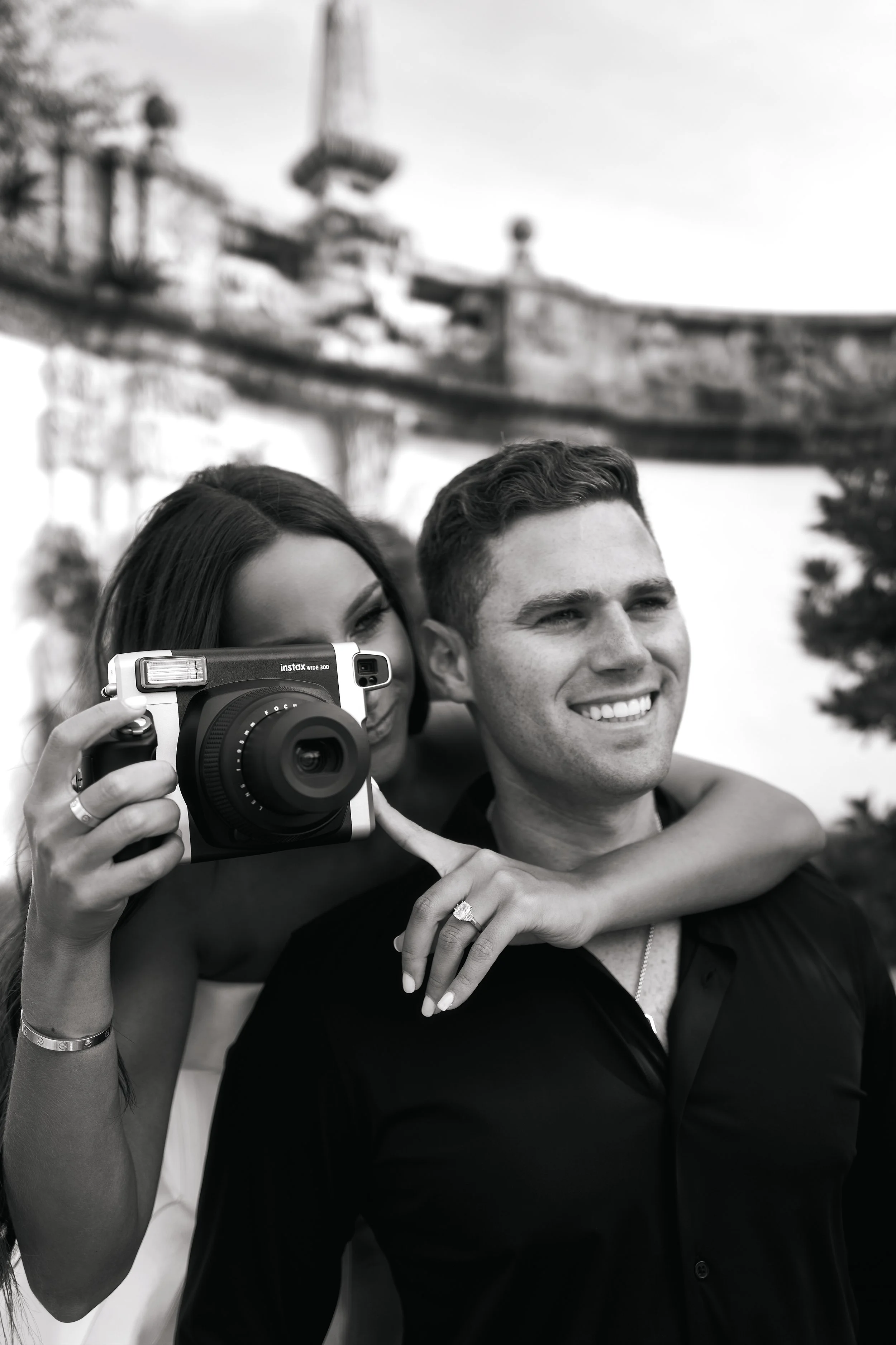 Black and white photo of a smiling man and woman taking a selfie together, with the woman holding a camera and the man hugging her from behind, at an amusement park with a roller coaster in the background.