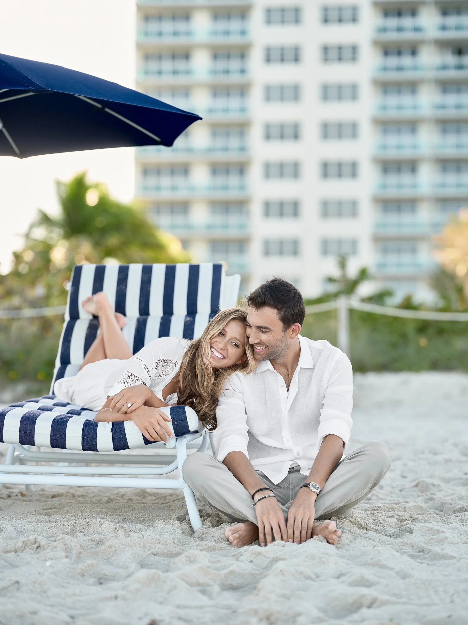 A happy couple smiling and lying on the sand at the beach, with a large building in the background, relaxing during daytime.