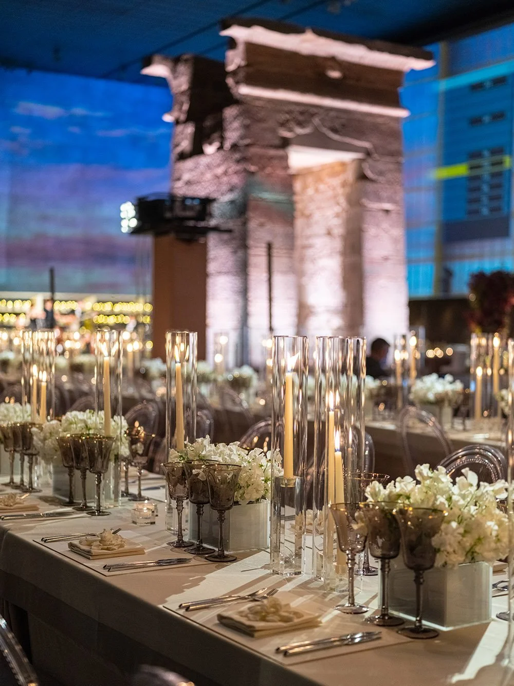 Elegant banquet table with white flowers, tall candles, and place settings, set up at an event with illuminated ancient ruins or stone structure in the background.