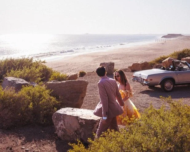 A couple holding hands near rocks and bushes on a beach with an ocean and a clear sky in the background, next to a vintage convertible car.