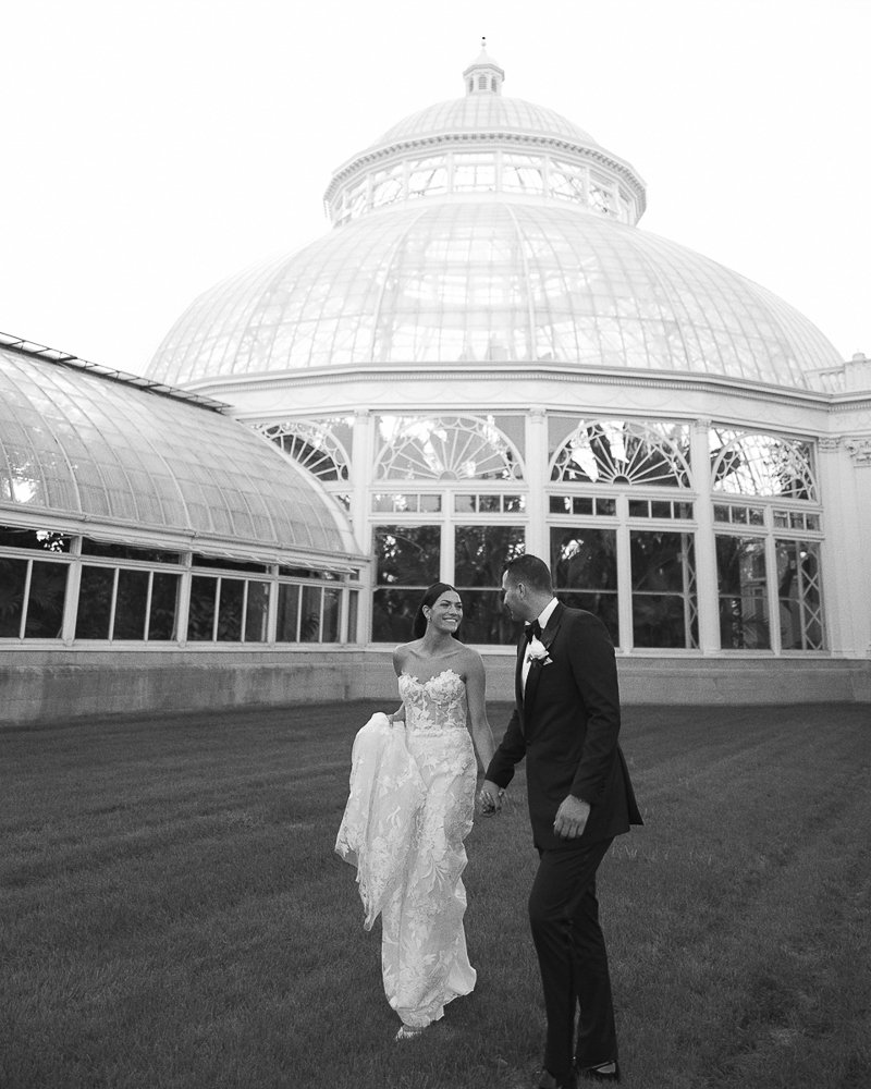 A bride and groom holding hands and looking at each other in front of a large glass conservatory or greenhouse, with a domed roof, in black and white.