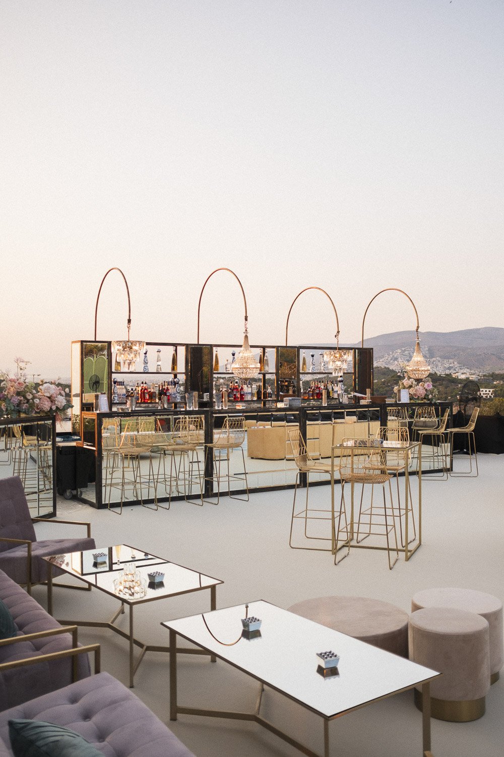Elegant rooftop bar with gold furniture, chandeliers, floral arrangements, and a distant view of mountains during sunset.