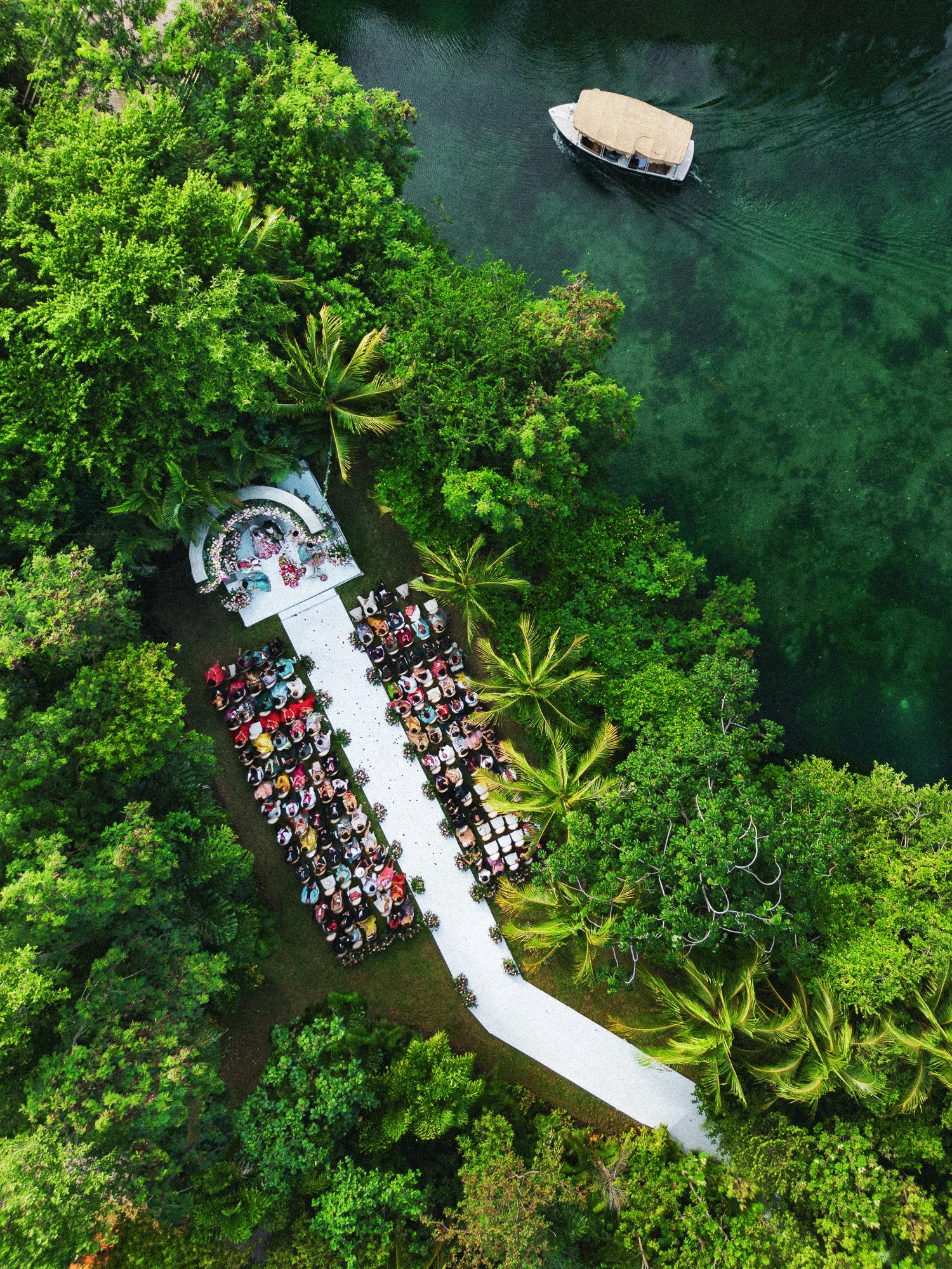 An aerial view of a boat on a river near a lush green forest, with rows of chairs and a small stage set up along a path.