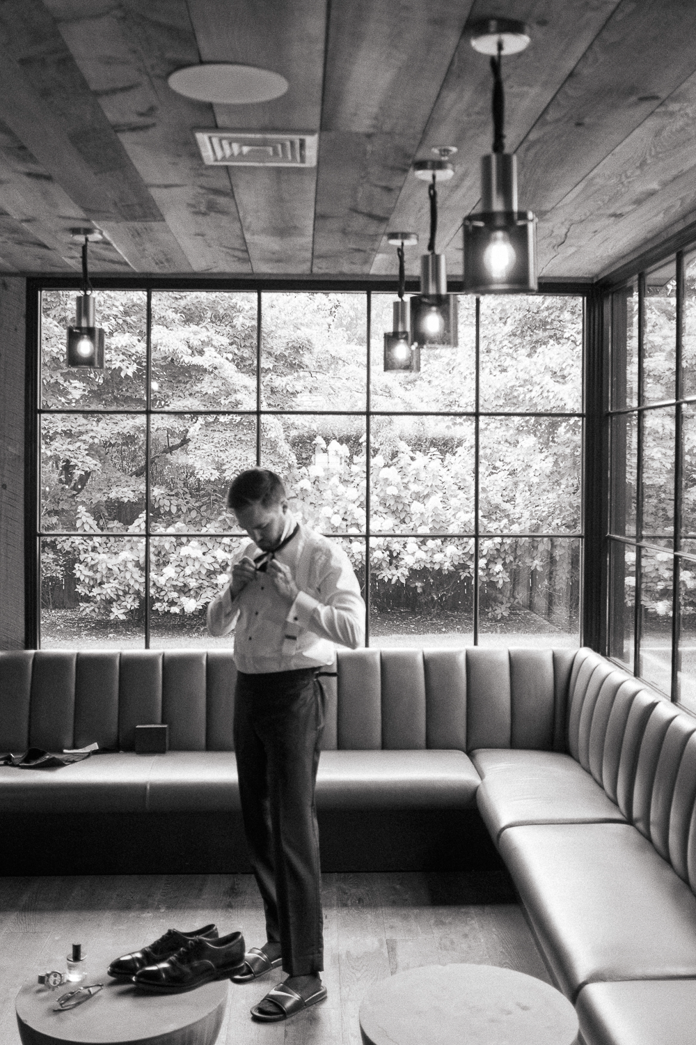 A man in formal attire adjusting his bow tie inside a modern, well-lit room with large windows and a garden view.