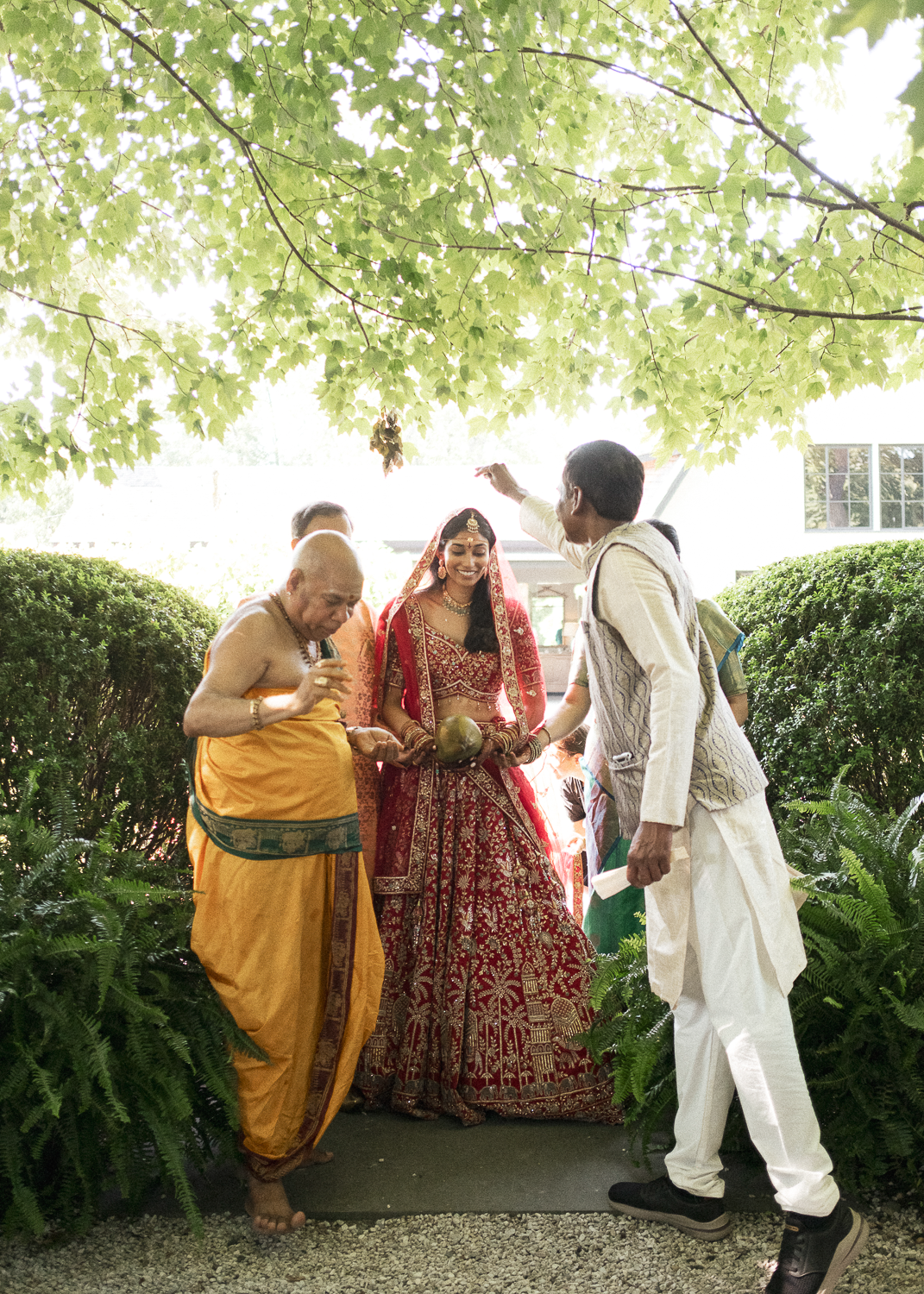 A wedding ceremony featuring a bride in a red and gold traditional Indian outfit, holding a coconut, standing under green leafy trees while people around her participate in rituals.