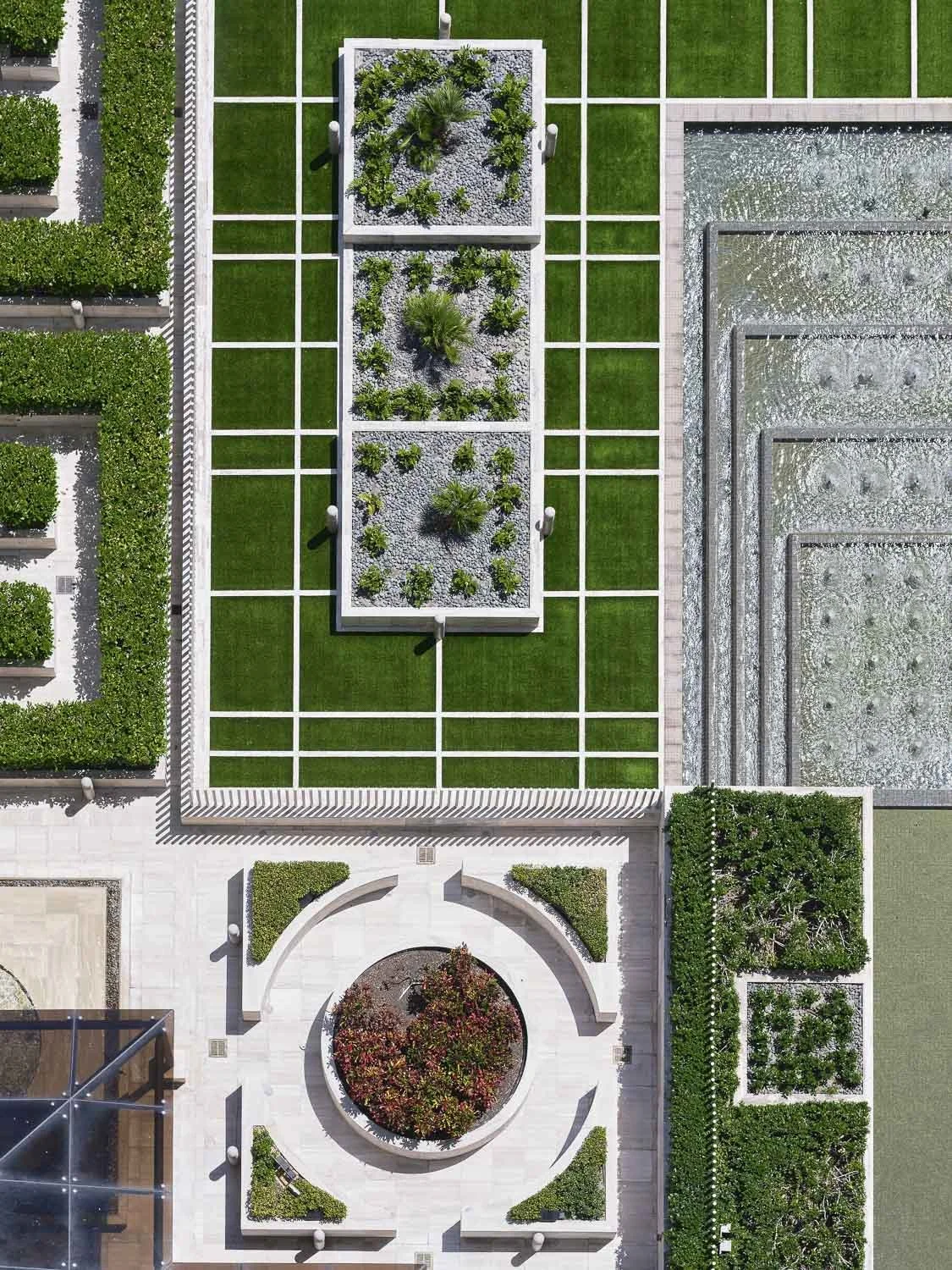 An aerial view of a landscaped garden with green lawns, rectangular garden beds with plants, water features, and paved walkways.