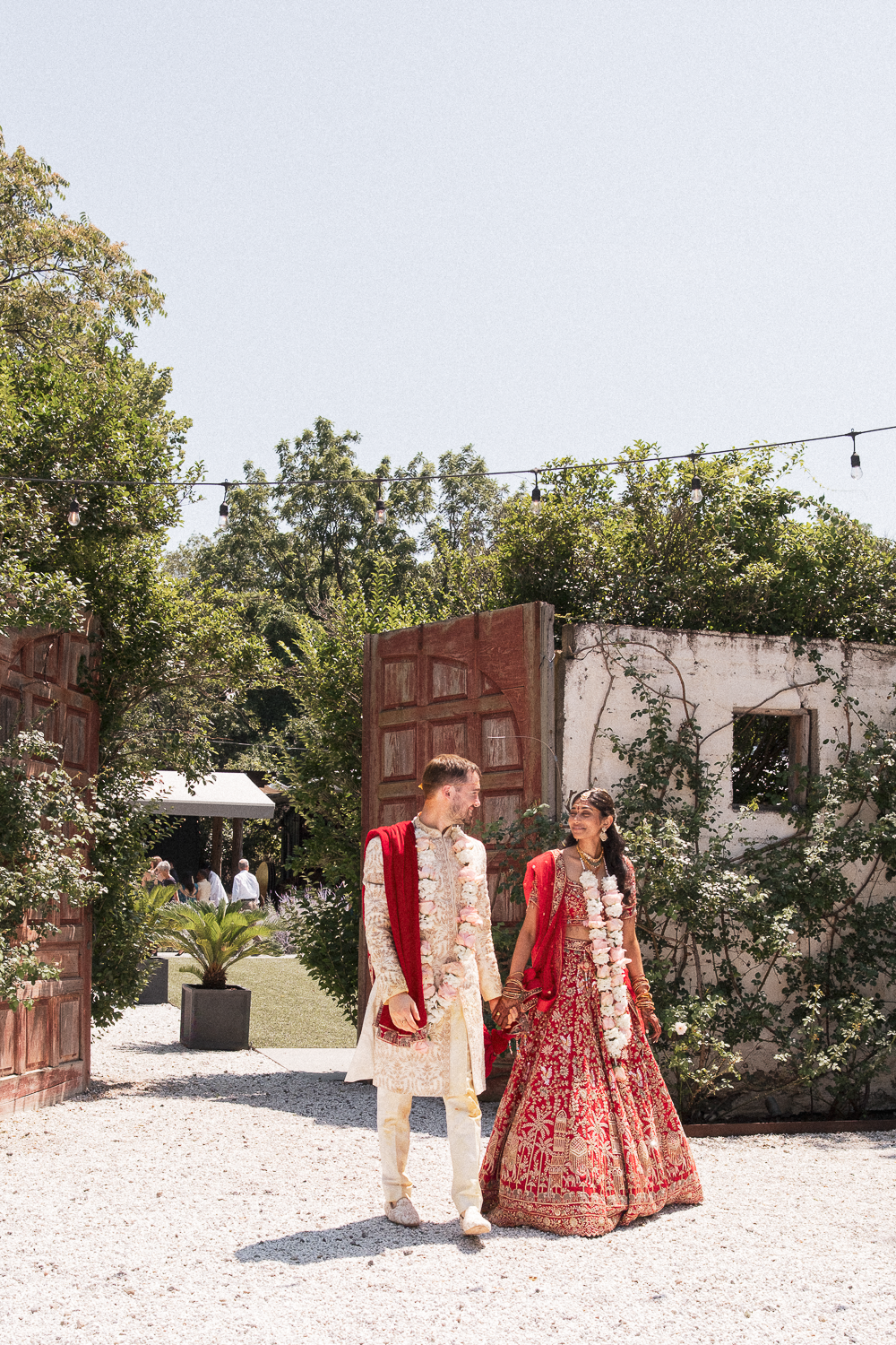 A couple dressed in traditional Indian wedding attire walking outdoors in a garden setting, holding hands, with decorative string lights overhead.