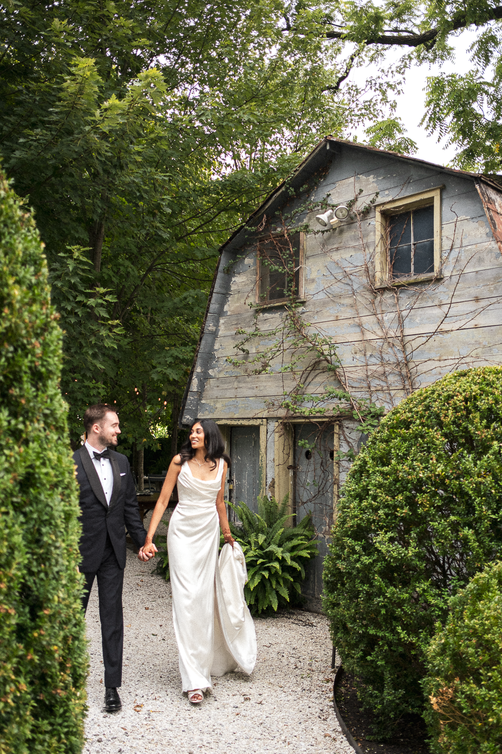 A couple dressed in wedding attire walking hand-in-hand along a gravel path beside an old, weathered house with vines growing on it. The man wears a black tuxedo and the woman wears a white wedding dress, smiling at each other.