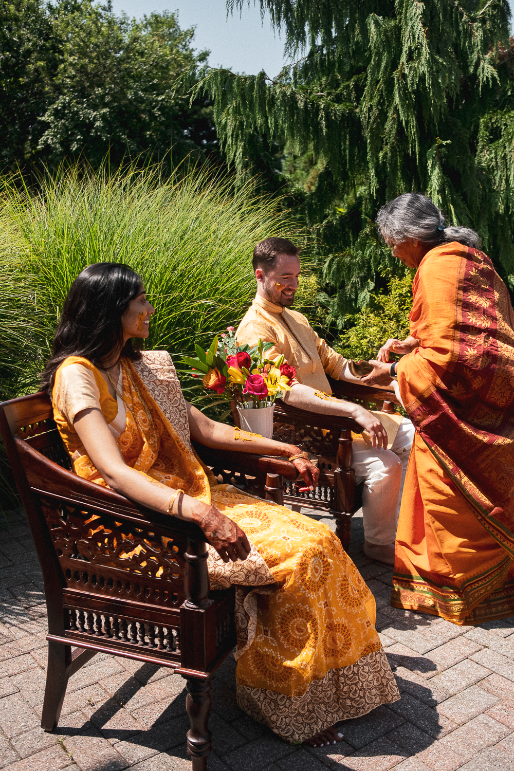 A multicultural couple dressed in traditional Indian attire sitting on wooden chairs outdoors, receiving a blessing from an older woman in Indian clothing during a wedding ceremony with lush greenery in the background.
