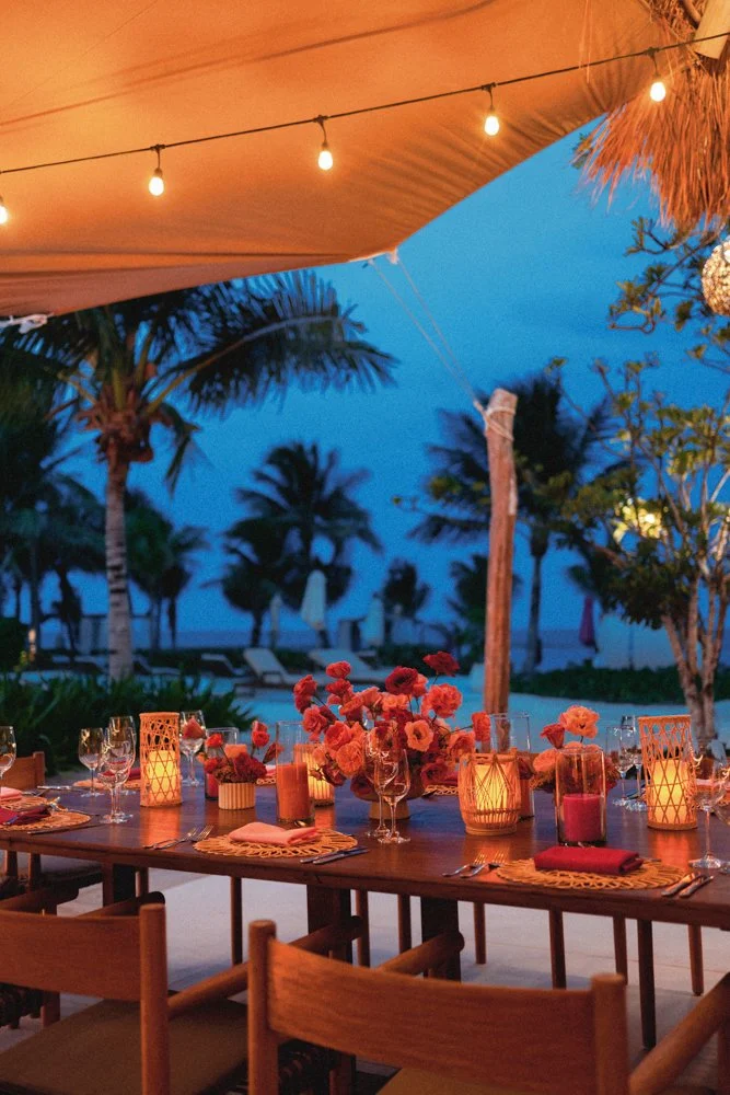 A dinner setup on a wooden table with pink and orange flowers, lit candles, and wine glasses, under string lights on a beachside patio with palm trees and lounge chairs in the background during twilight.