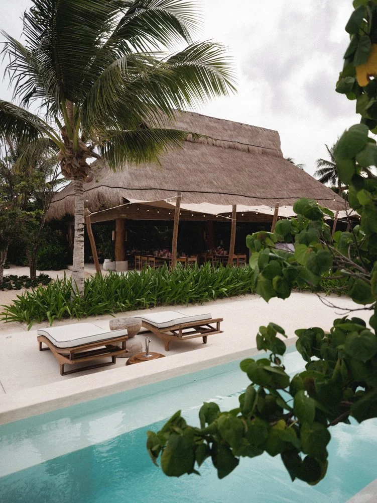 Poolside view at a tropical resort, featuring lounge chairs and a thatched-roof cabana surrounded by greenery and palm trees.