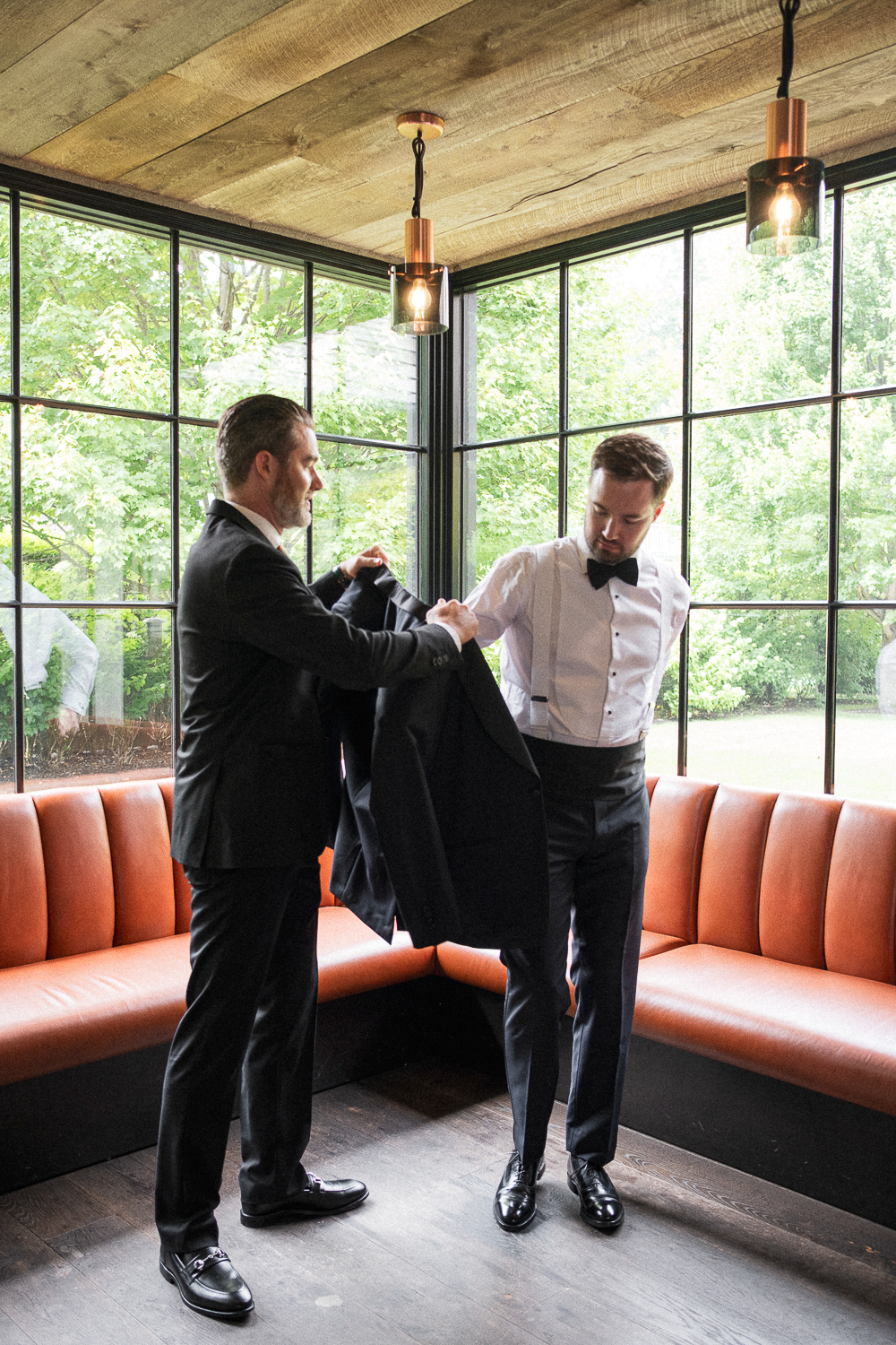 Two men in formal tuxedos, one helping the other with his jacket in a modern, bright room with large windows and greenery outside.