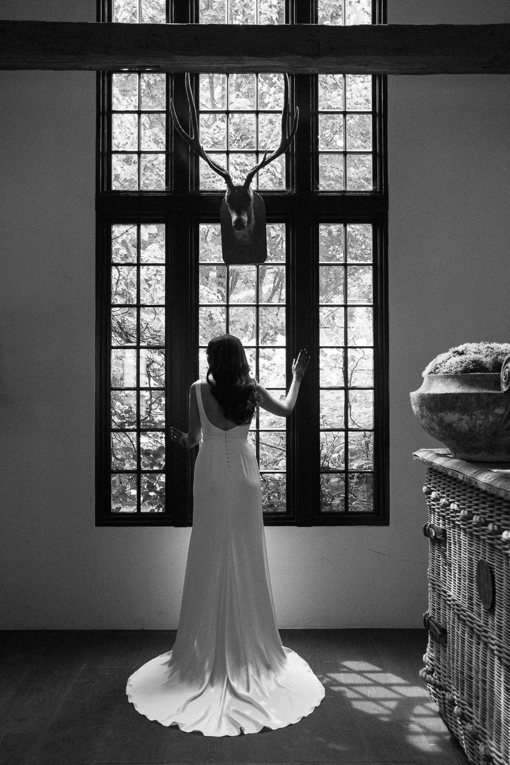 A woman in a long white dress standing indoors, facing a large window, with her back to the camera. A mounted deer head is visible above the window, and sunlight filters through the window onto the floor.
