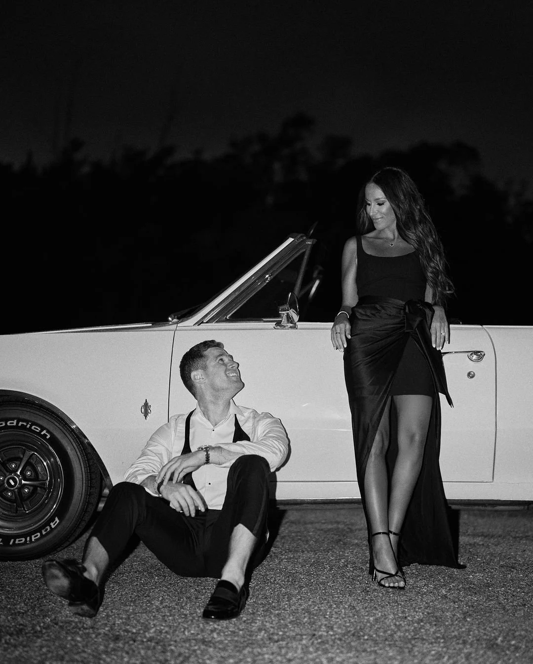A black-and-white photo features a woman in a dress standing beside a classic car, looking at a man sitting on the ground. The man is smiling up at her, dressed in a white shirt and dark pants, with his shoes off and legs crossed. The setting appears