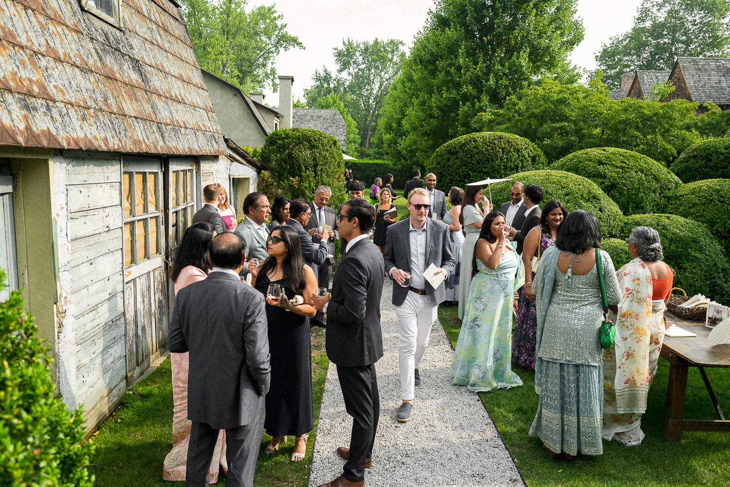 A group of people dressed in formal and semi-formal attire mingling outdoors at a garden party on a sunny day. Some are holding drinks, and there are tables with items on them to the right.