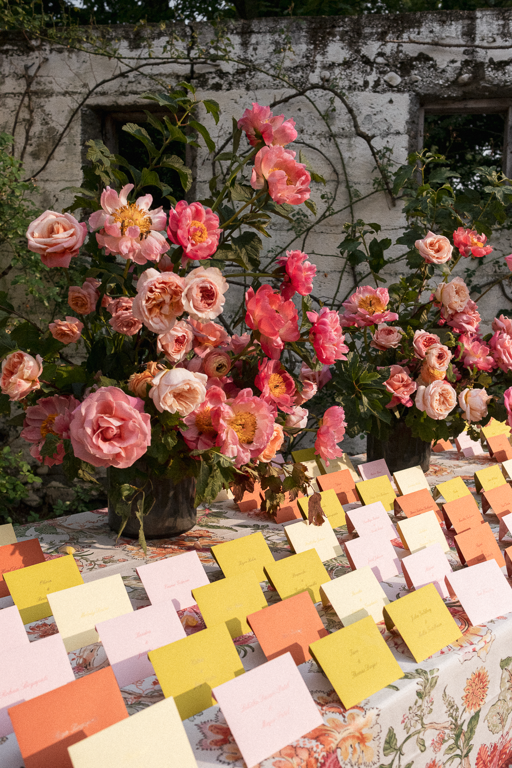 Pink and peach roses arranged in black vases on a floral tablecloth, with colorful place cards nearby, against a rustic wall background.