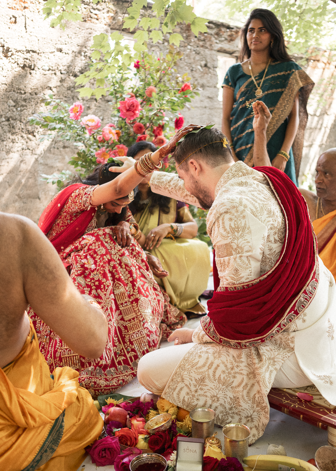 Indian wedding ceremony with bride and groom dressed in traditional attire, participating in a ritual, surrounded by family and friends, with flowers and ritual items on the ground.