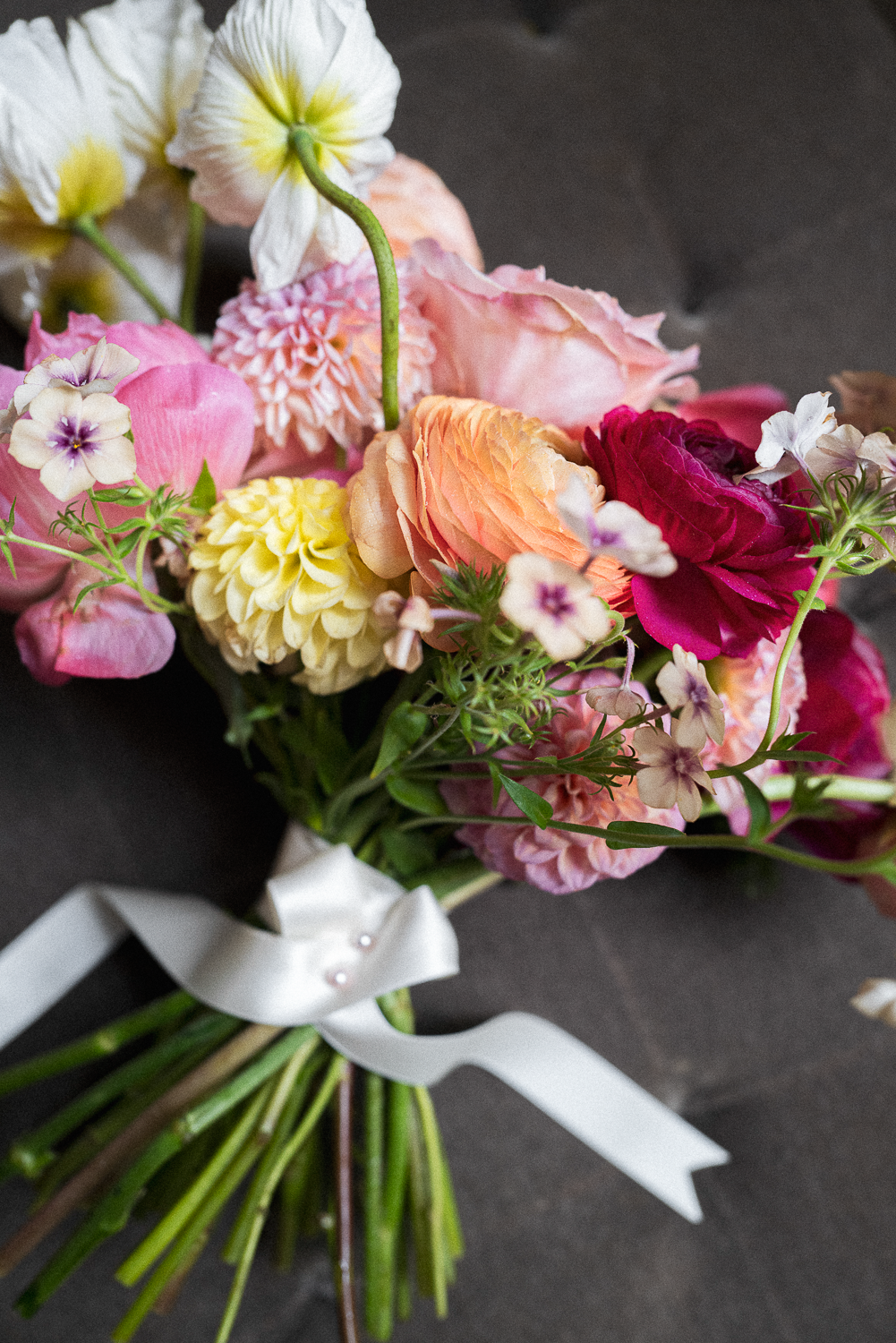 Colorful bouquet of various flowers including roses, statice, and ranunculus tied with a white ribbon on a dark surface.