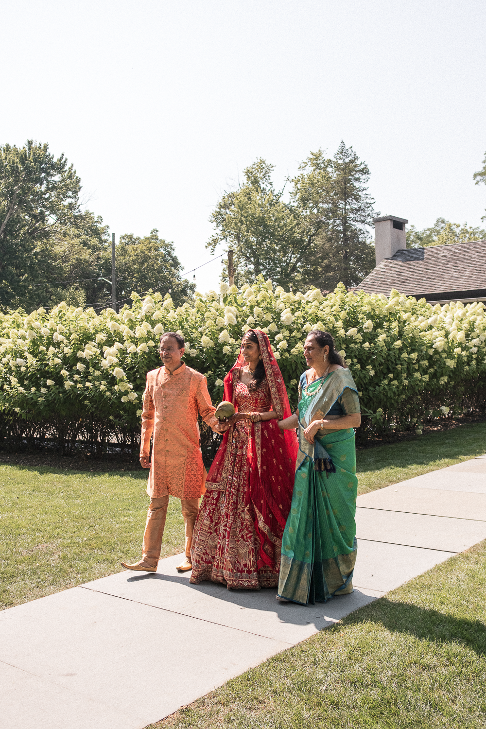 Three people walking outdoors on a cement path, dressed in traditional Indian attire, with a background of hydrangea bushes and a house with a chimney.
