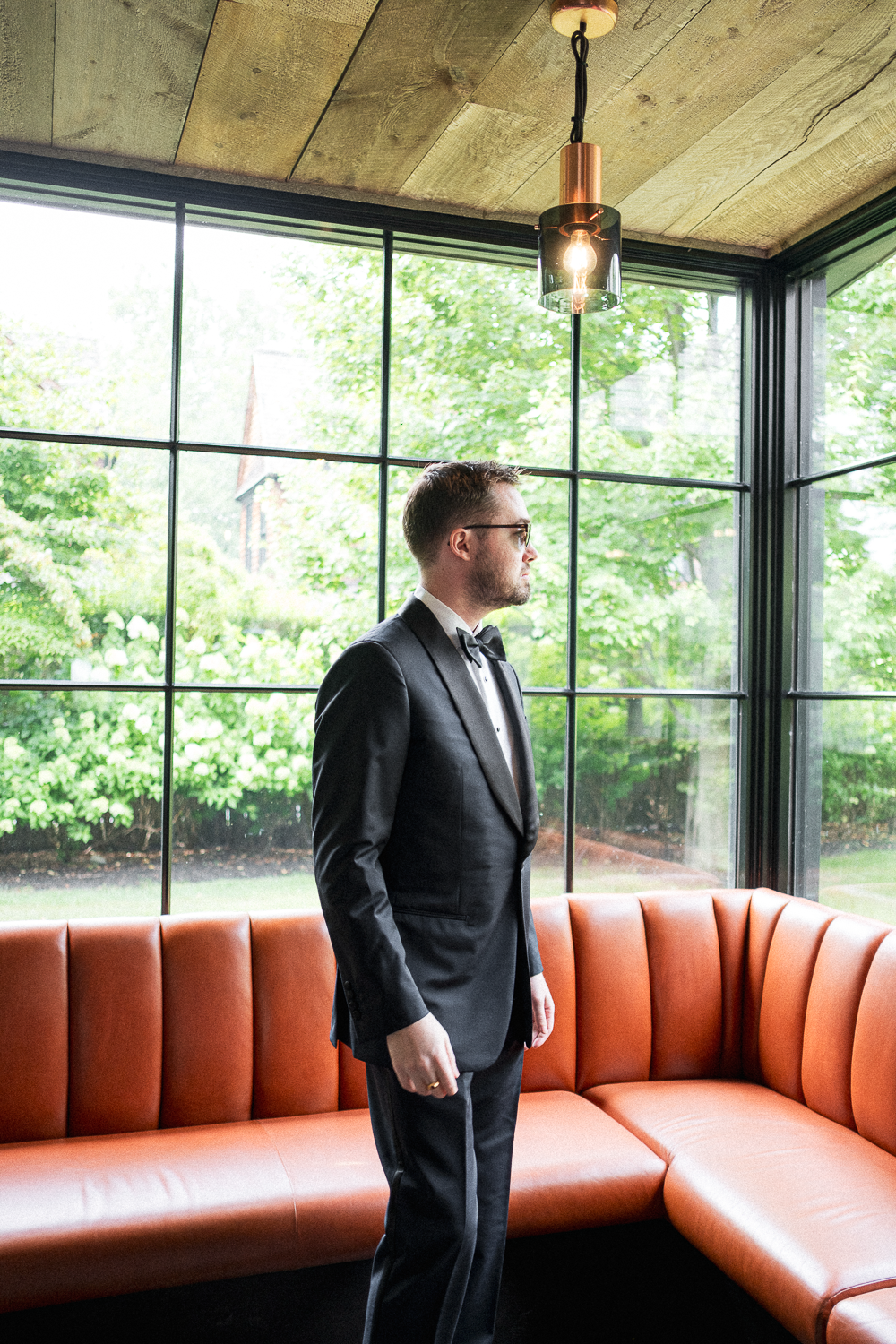 A man in a formal tuxedo with a bow tie and glasses standing indoors near a large window, with greenery outside, in front of an orange sectional sofa and a modern ceiling light.