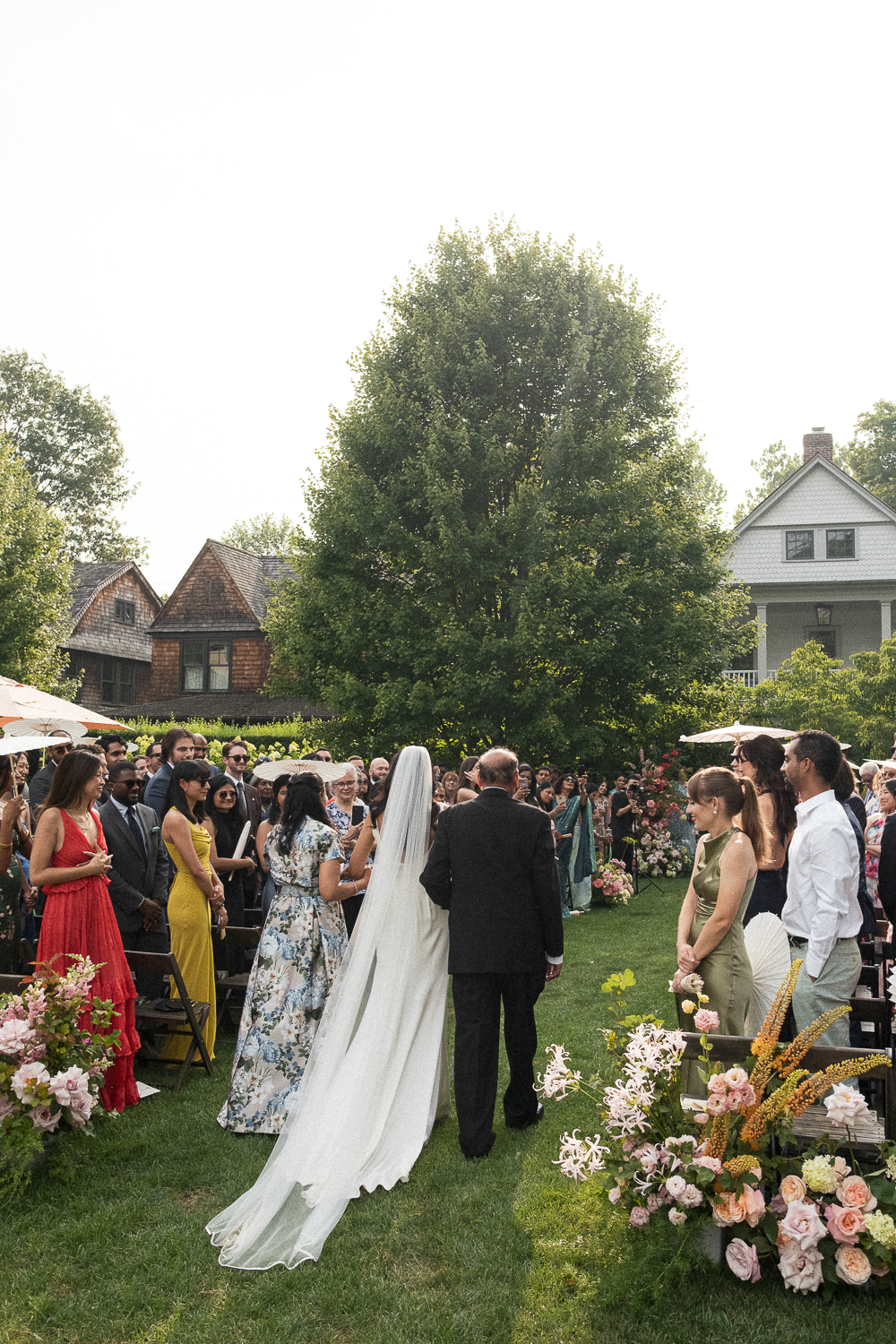 A bride in a white wedding gown with a long veil walking down the aisle with a man in a black suit at an outdoor wedding ceremony. Guests are standing on both sides, some holding umbrellas, with flowers and greenery around.