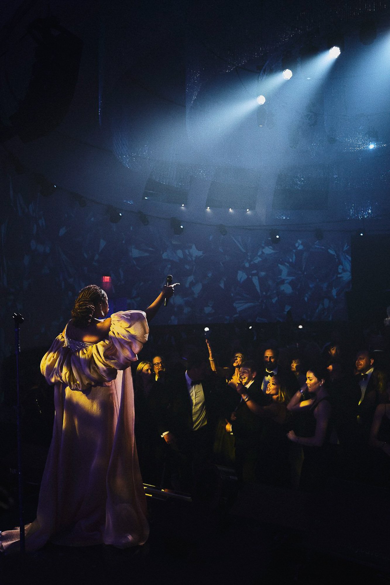 A performer on stage, wearing a flowing, elegant dress, holding a microphone, engaging with a crowd in a dimly lit venue with blue stage lighting and an audience in formal attire.
