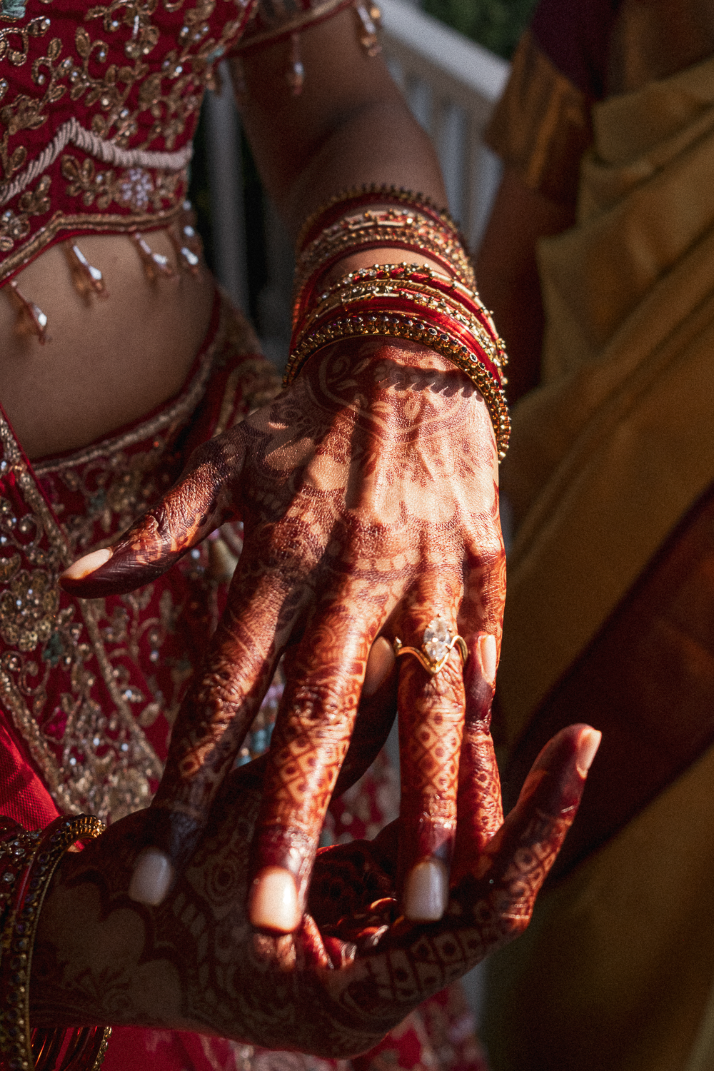 Close-up of a woman's decorated hand and wrist with henna designs, gold jewelry, and red bangles, wearing traditional vibrant red and gold embroidered attire.