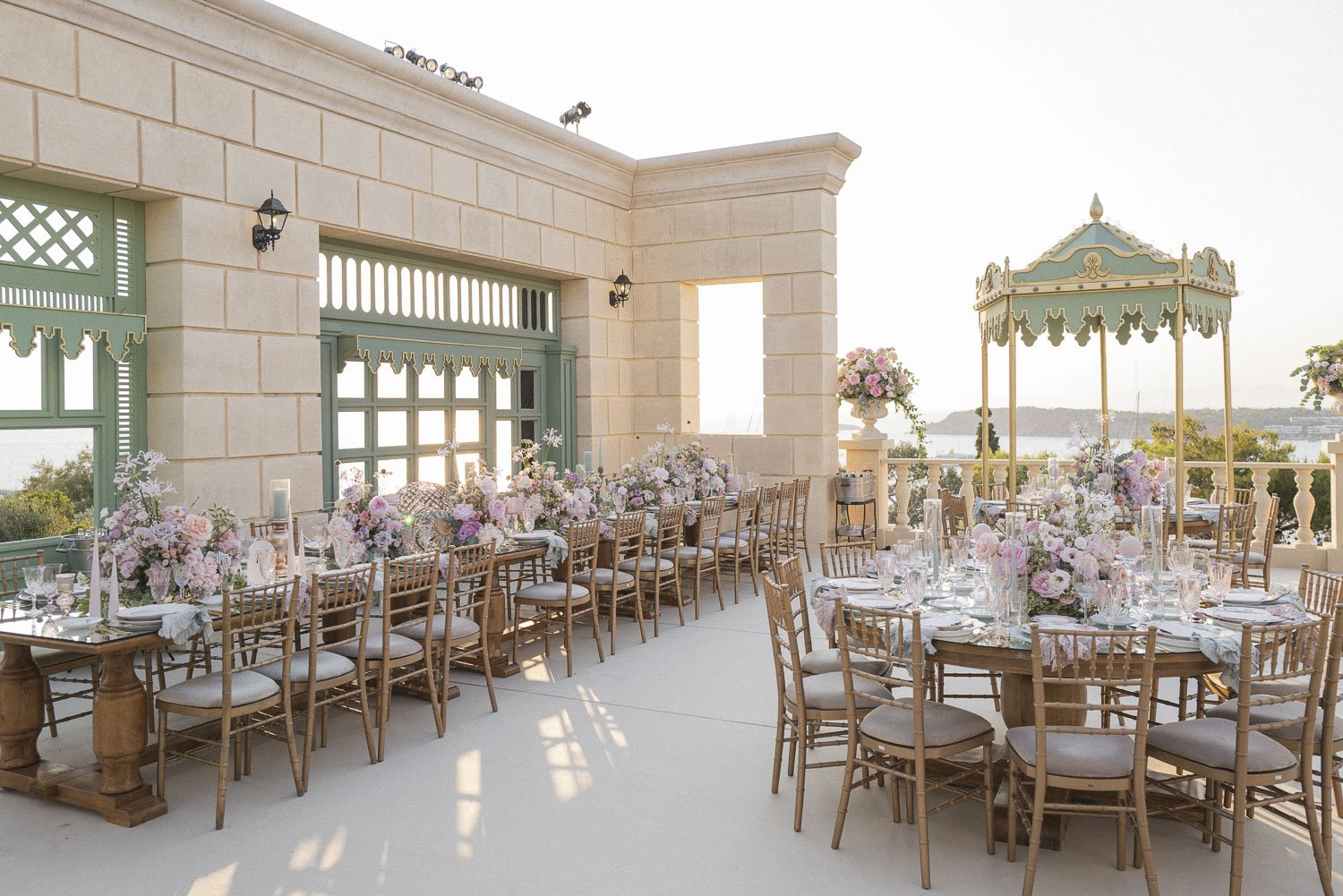 Elegant outdoor wedding reception setup with round tables, floral centerpieces, Chiavari chairs, and a vintage-style gazebo with a view of water and landscape in the background.
