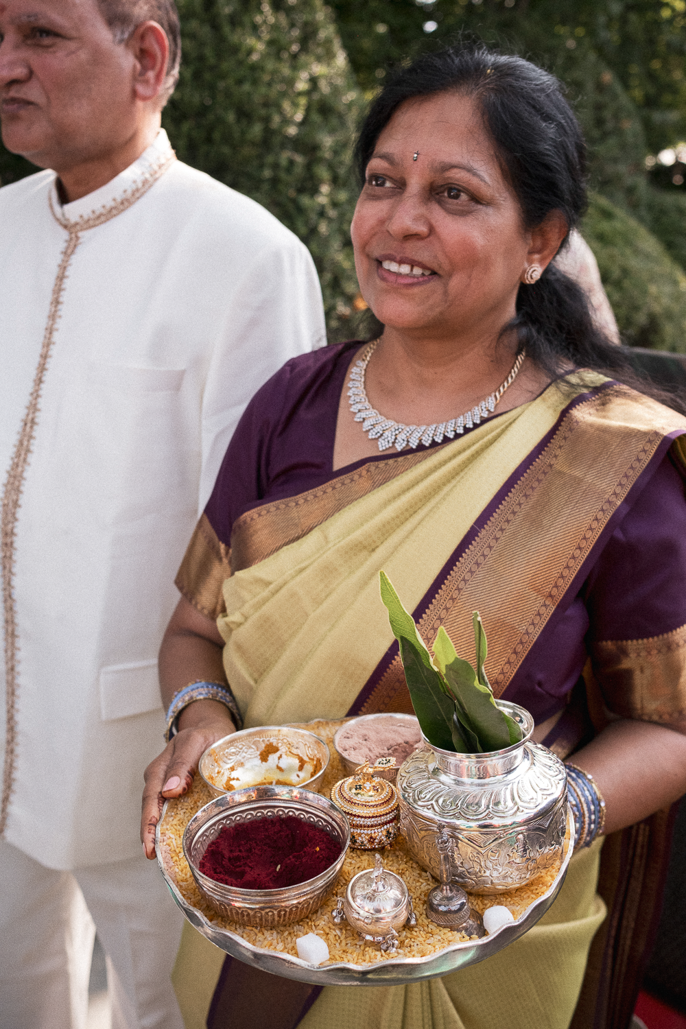 An Indian woman dressed in a purple and gold sari holding a silver tray with traditional ceremonial items, including a vase with green leaves, bowls of red powder, and decorative containers, during a cultural or religious celebration.