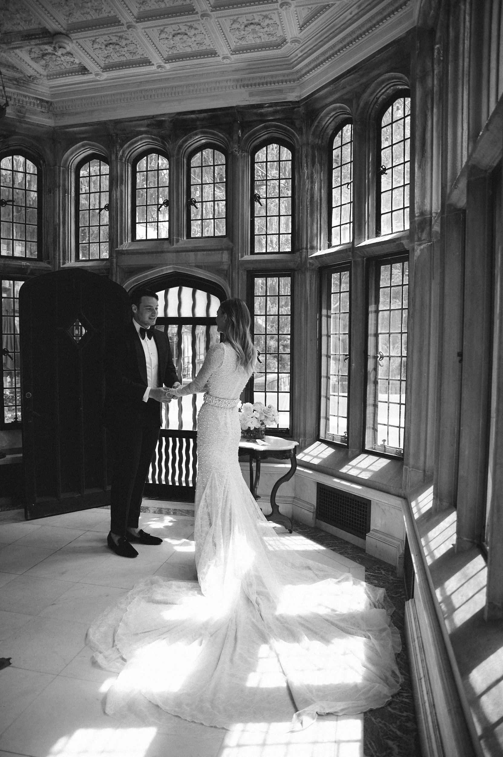 A bride and groom holding hands and smiling at each other during their wedding ceremony in a room with large windows and ornate ceiling details, with sunlight streaming in.